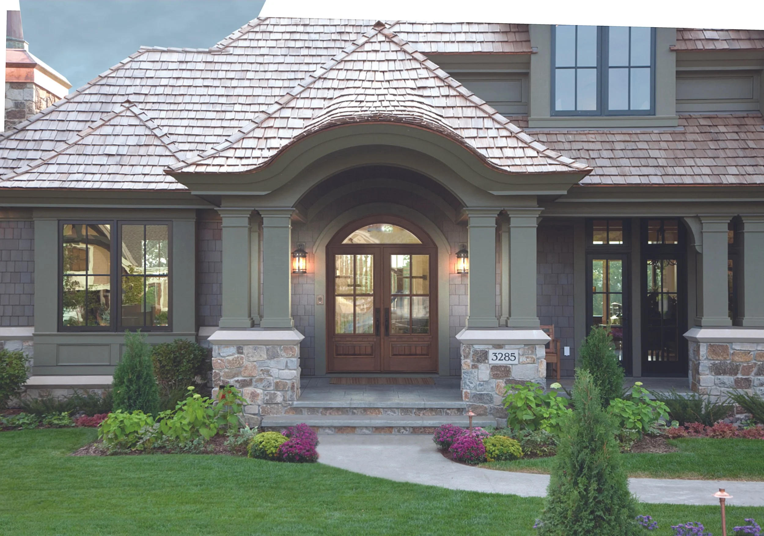 Front view of a modern house with a tiled roof, stone pillars, a wooden front door, and a well-kept lawn with plants and flowers.