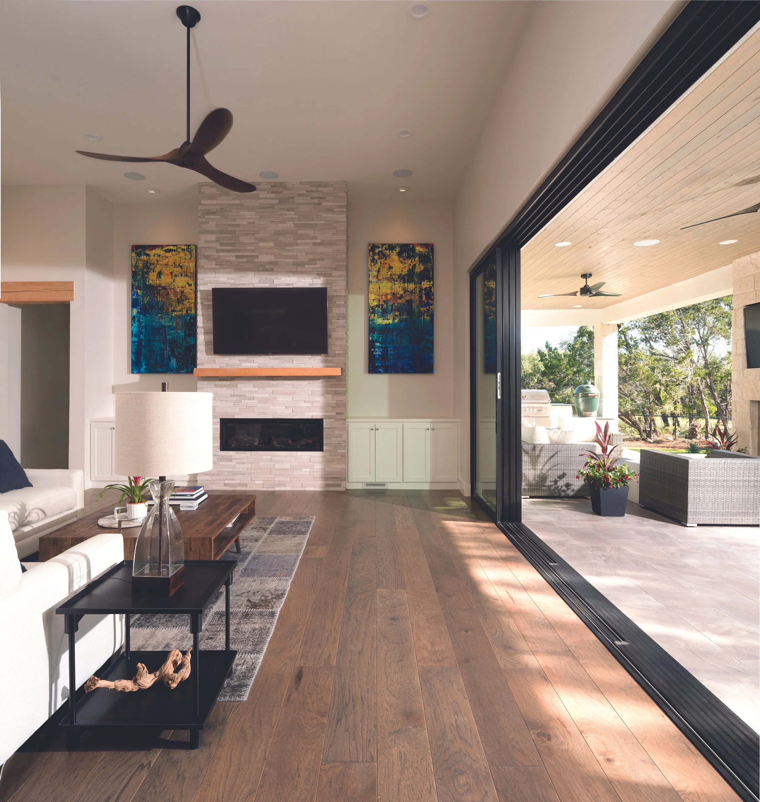 Living room with a fireplace, two colorful abstract paintings, a TV, a ceiling fan, a coffee table, a white sofa, and a sliding glass door leading to an outdoor patio with outdoor furniture and a view of trees.
