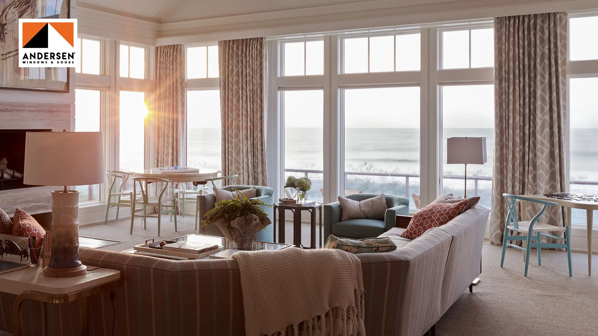 Living room with large windows showing ocean view, beige curtains, sofa, armchairs, coffee table, side tables, lamps, and a dining area in the background.