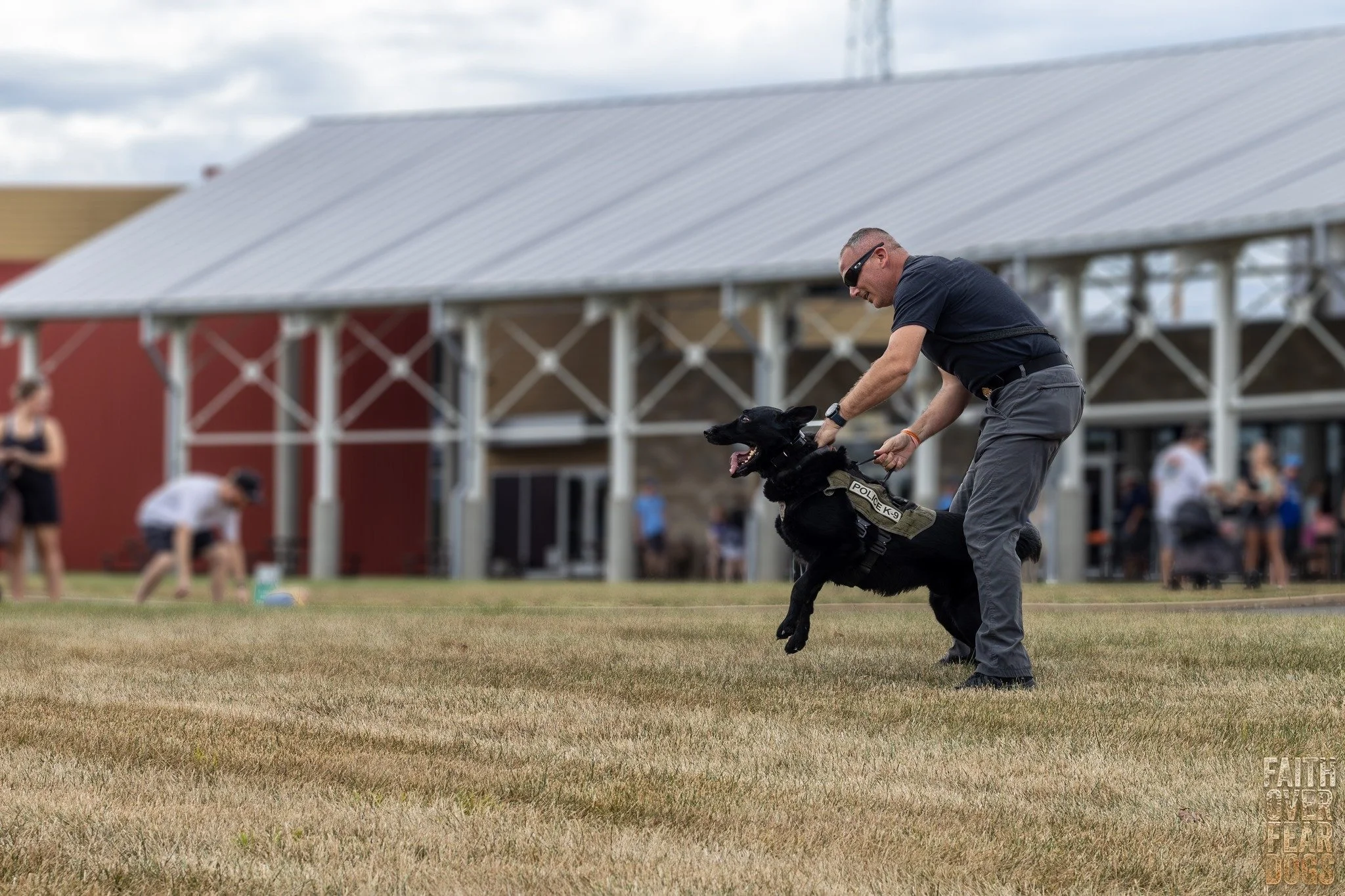 A man in sunglasses and a dark shirt, holding a police dog by the collar, practicing or performing on a grassy field with people and a building in the background.