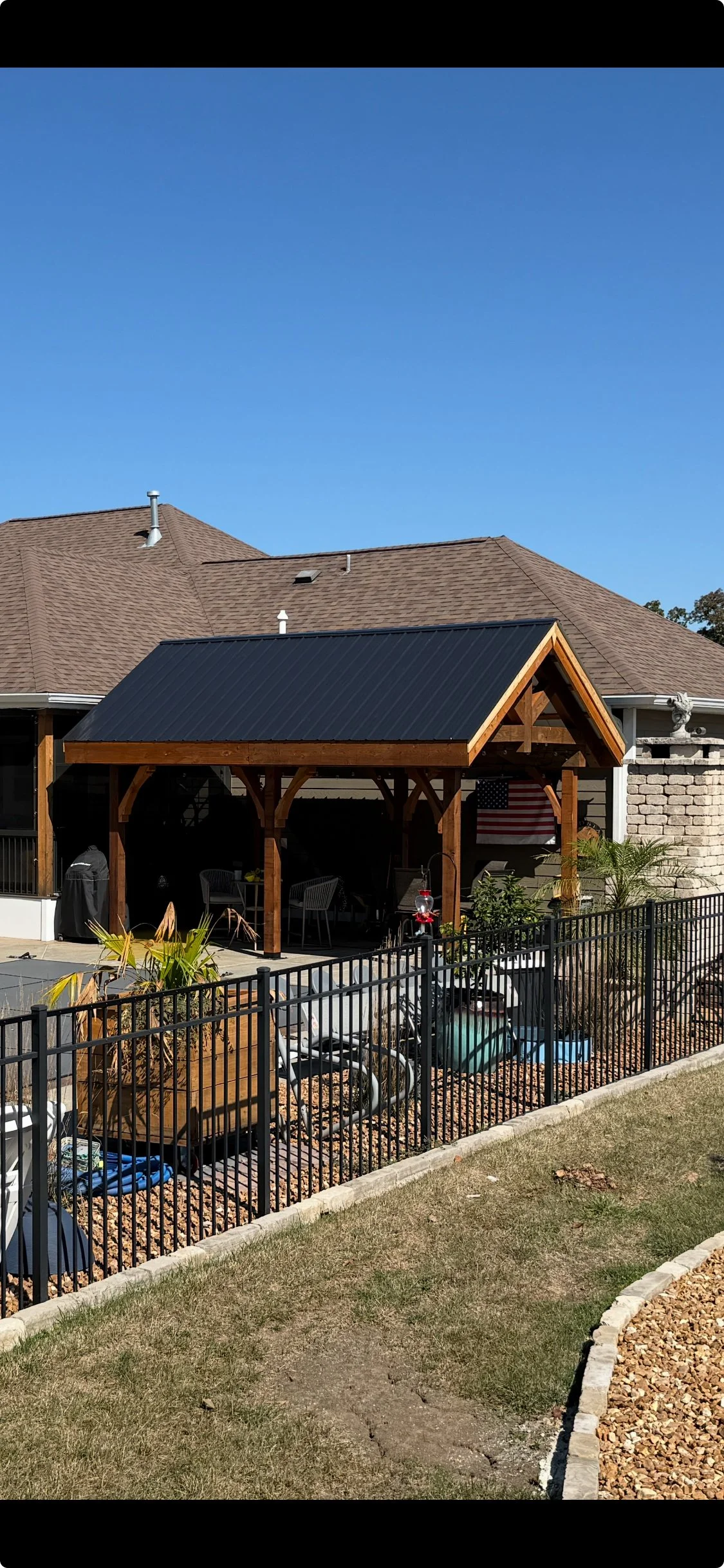 A backyard patio with a wooden pergola and black metal roof, outdoor furniture, a flag, and various plants and garden decorations, separated by a black metal fence from a grassy yard.
