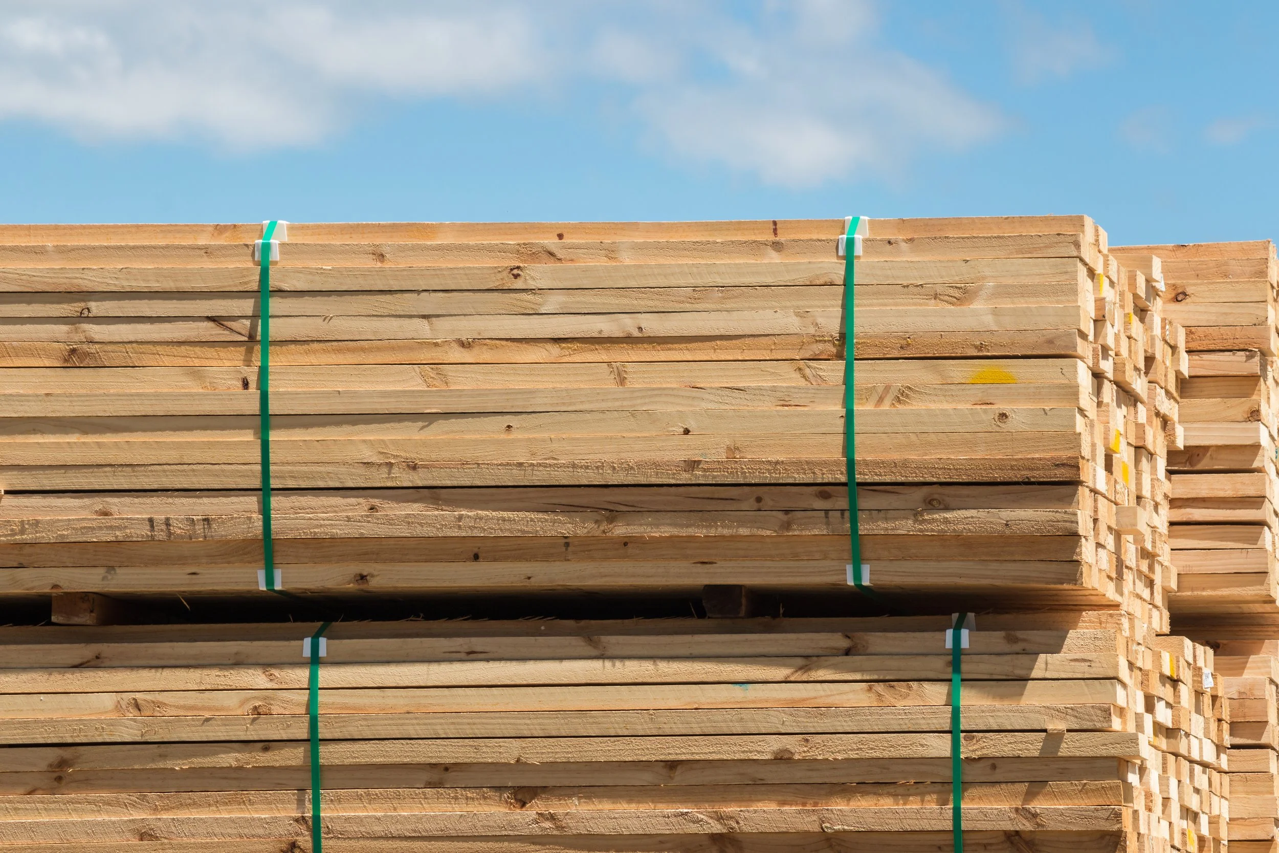 Stacked wooden planks secured with green straps under a blue sky with clouds.