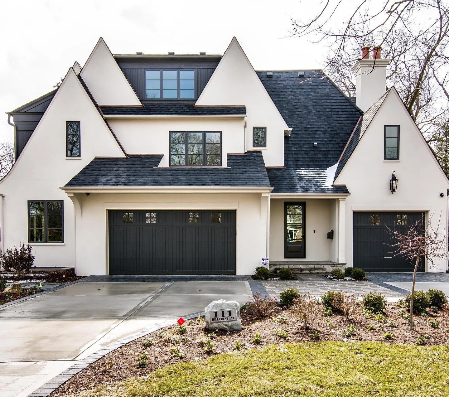 Modern white house with black garage doors and roof, multiple peaked roofs, large front windows, a small front yard, and a stone pathway leading to the front door.