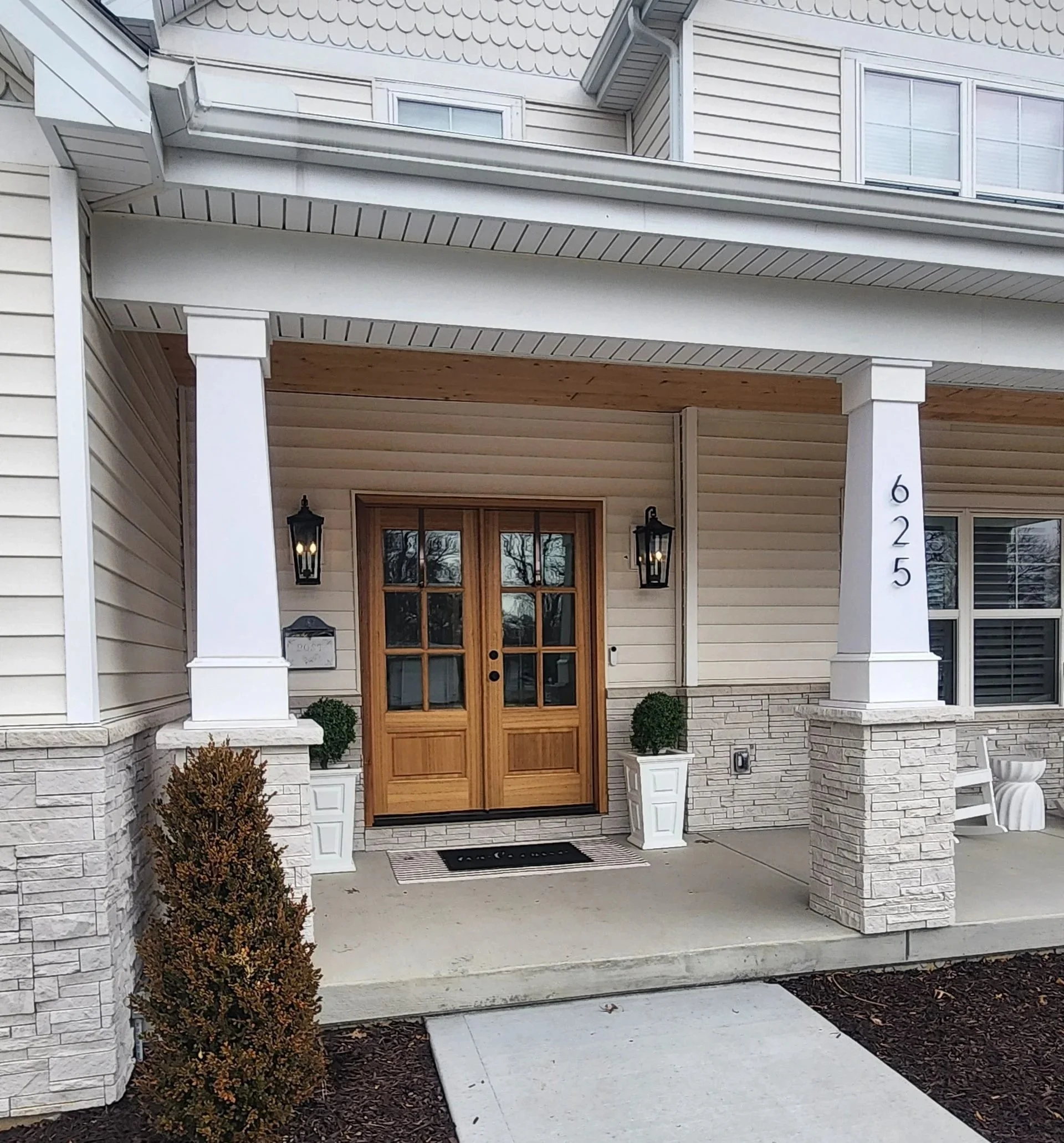 Front porch of a house with a wooden door, black lantern lights, two white planters with green bushes, and house number 625 on a white pillar.