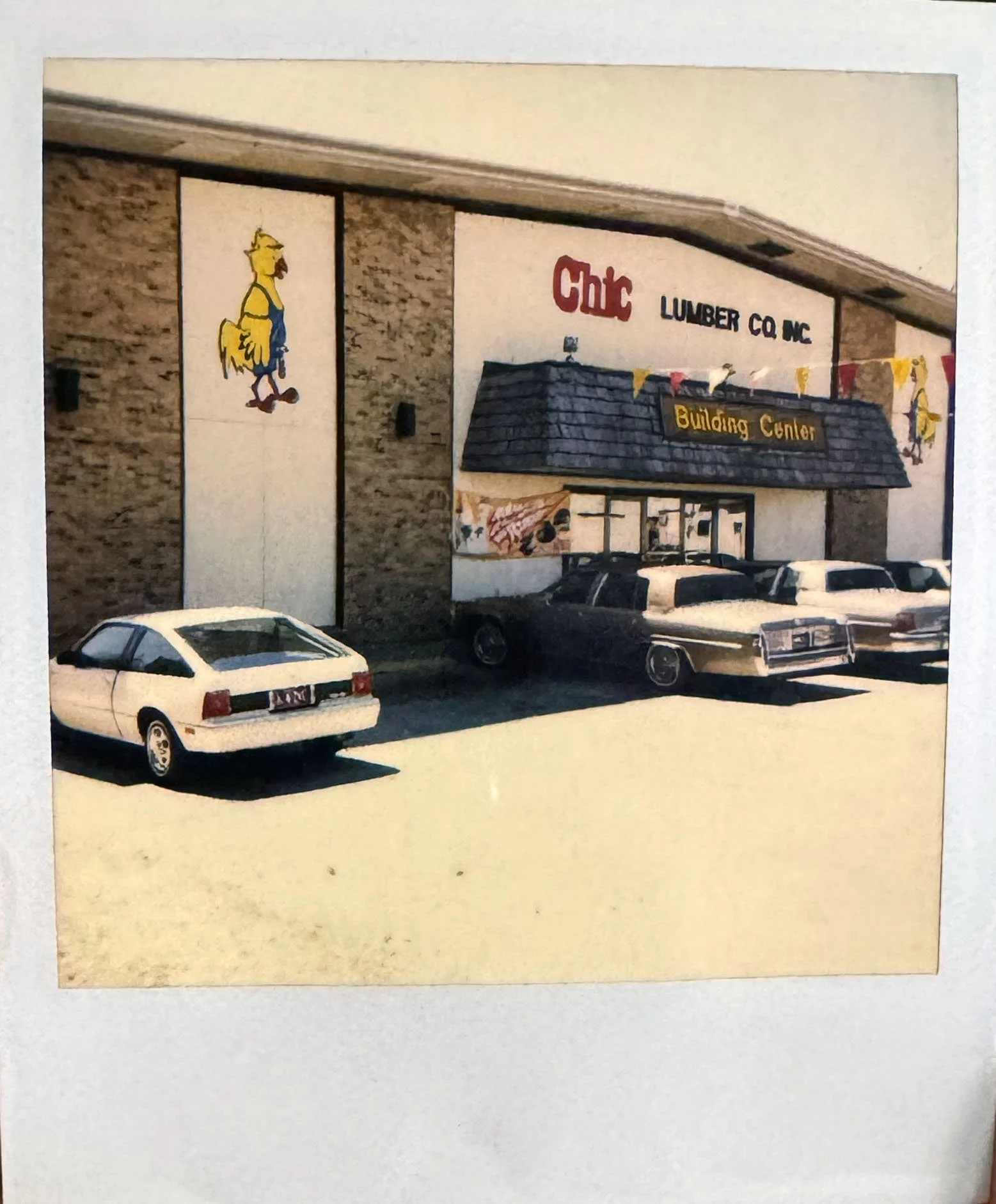 A building with signs reading "Chic Lumber Co. Inc" and "Building Center", decorated with yellow and red pennant banners, and surrounded by parked cars.