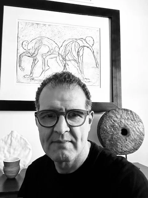 A man with glasses taking a selfie in front of a framed abstract line drawing of two figures with bent knees, on a wall. Behind him, there is a round textured sculpture and some pottery.