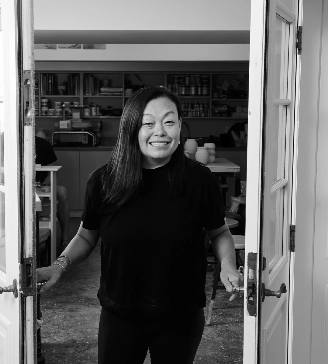 A woman smiling while standing in a doorway, with a kitchen or workshop visible in the background.