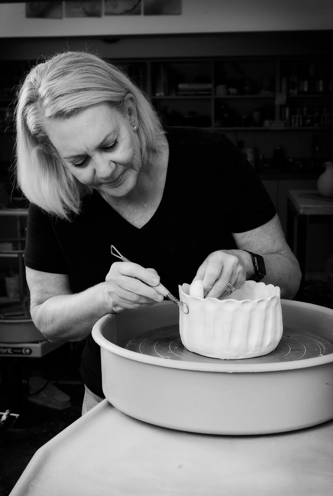 A woman is working on a ceramic piece in a pottery studio, using a tool to add details to a wavy-edged vessel on a spinning pottery wheel.