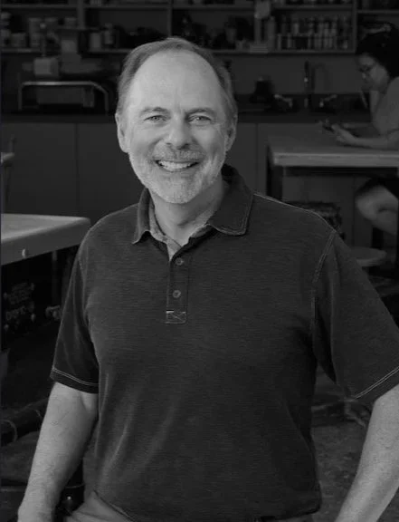 A smiling middle-aged man with a beard and short hair, wearing a polo shirt, standing in a laboratory or workshop setting.
