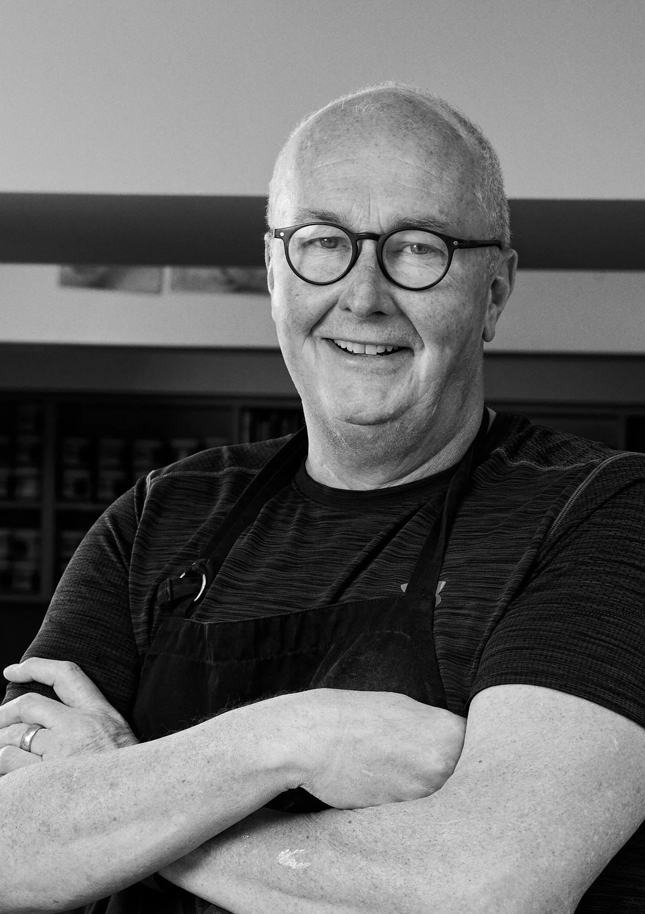 Black-and-white photo of a smiling man with glasses wearing a dark shirt and apron, crossing his arms, standing in what appears to be a kitchen or bookstore background.