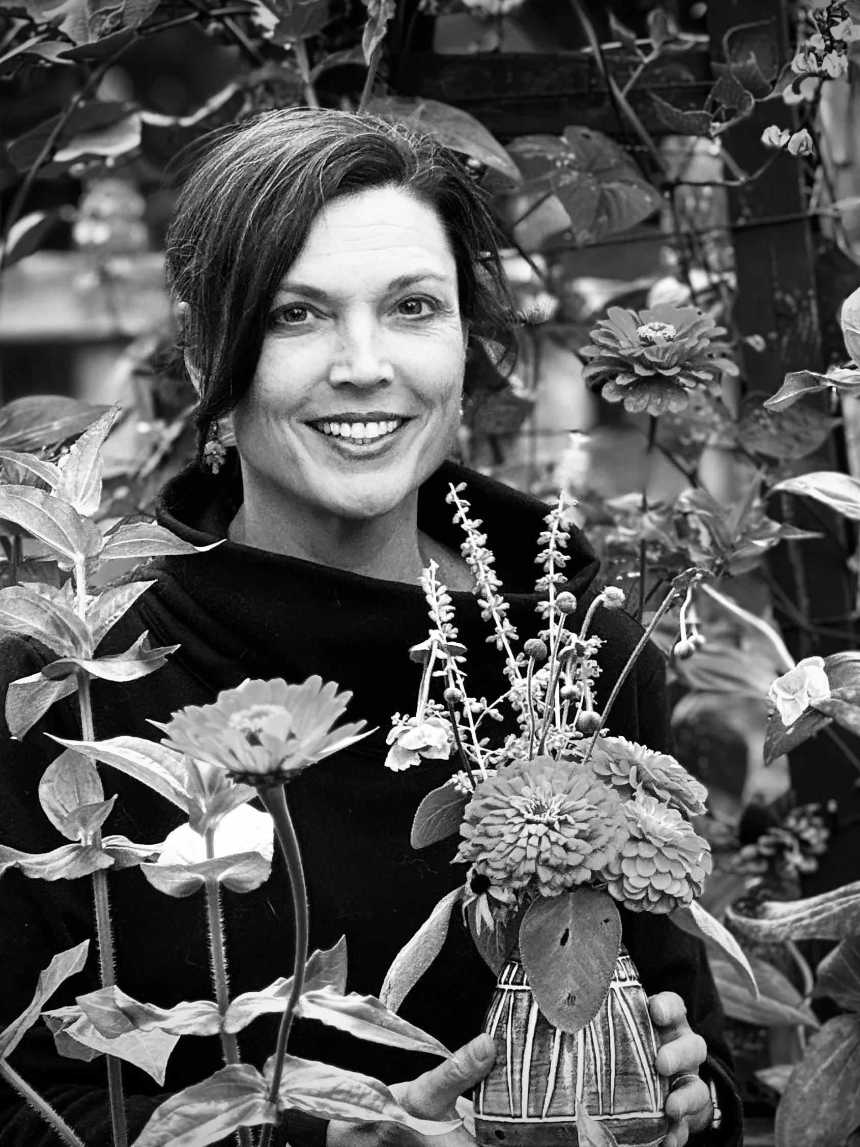 A woman with short dark hair smiling, holding a striped ceramic pot filled with flowers, surrounded by lush foliage in a garden setting.