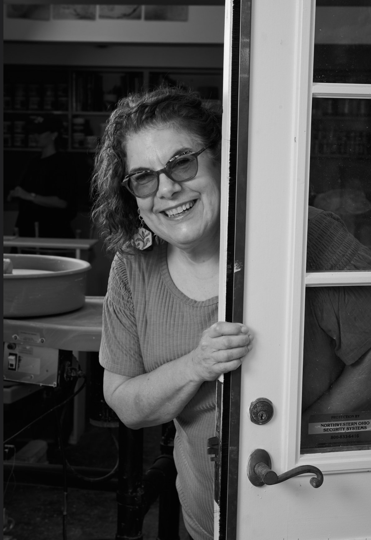 A woman with curly hair and glasses is smiling and peeking through a partly open door in a room that appears to be a kitchen or workshop.
