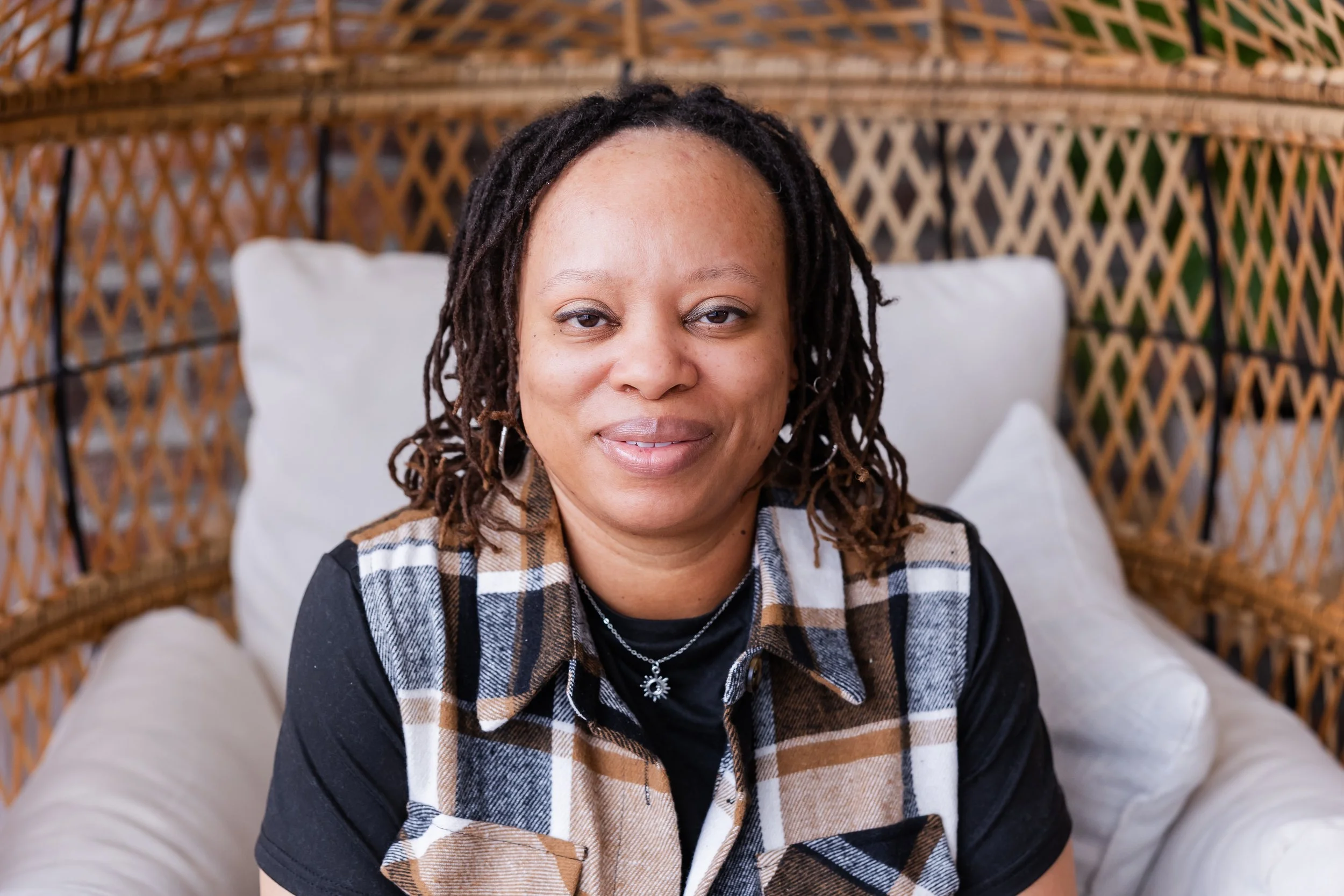 A woman with shoulder-length dreadlocks, wearing a black t-shirt and a plaid sleeveless shirt, sitting on a porch with wicker walls and white cushions.
