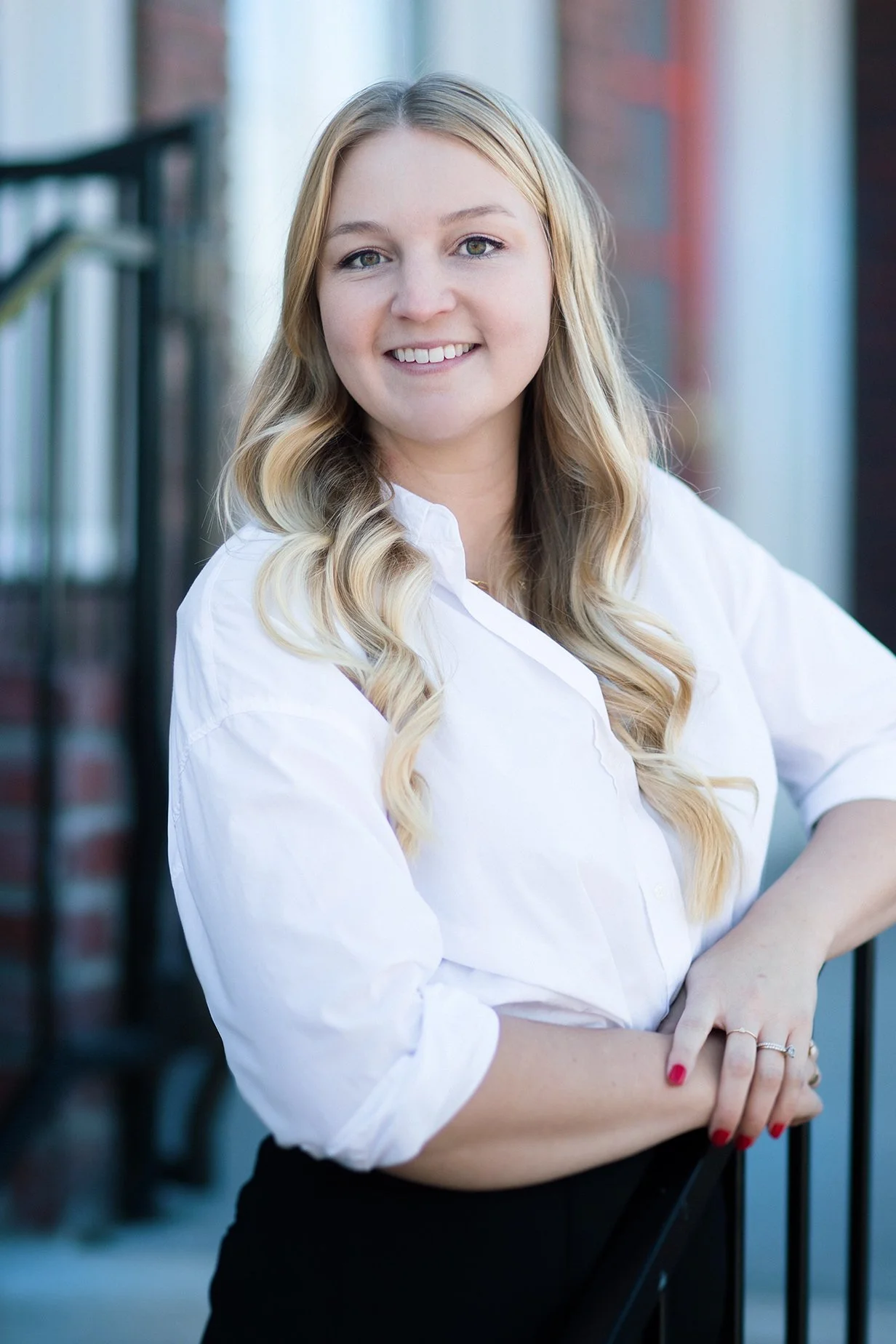 A woman with long blonde wavy hair, wearing a white shirt, smiling and leaning on a balcony railing outdoors.
