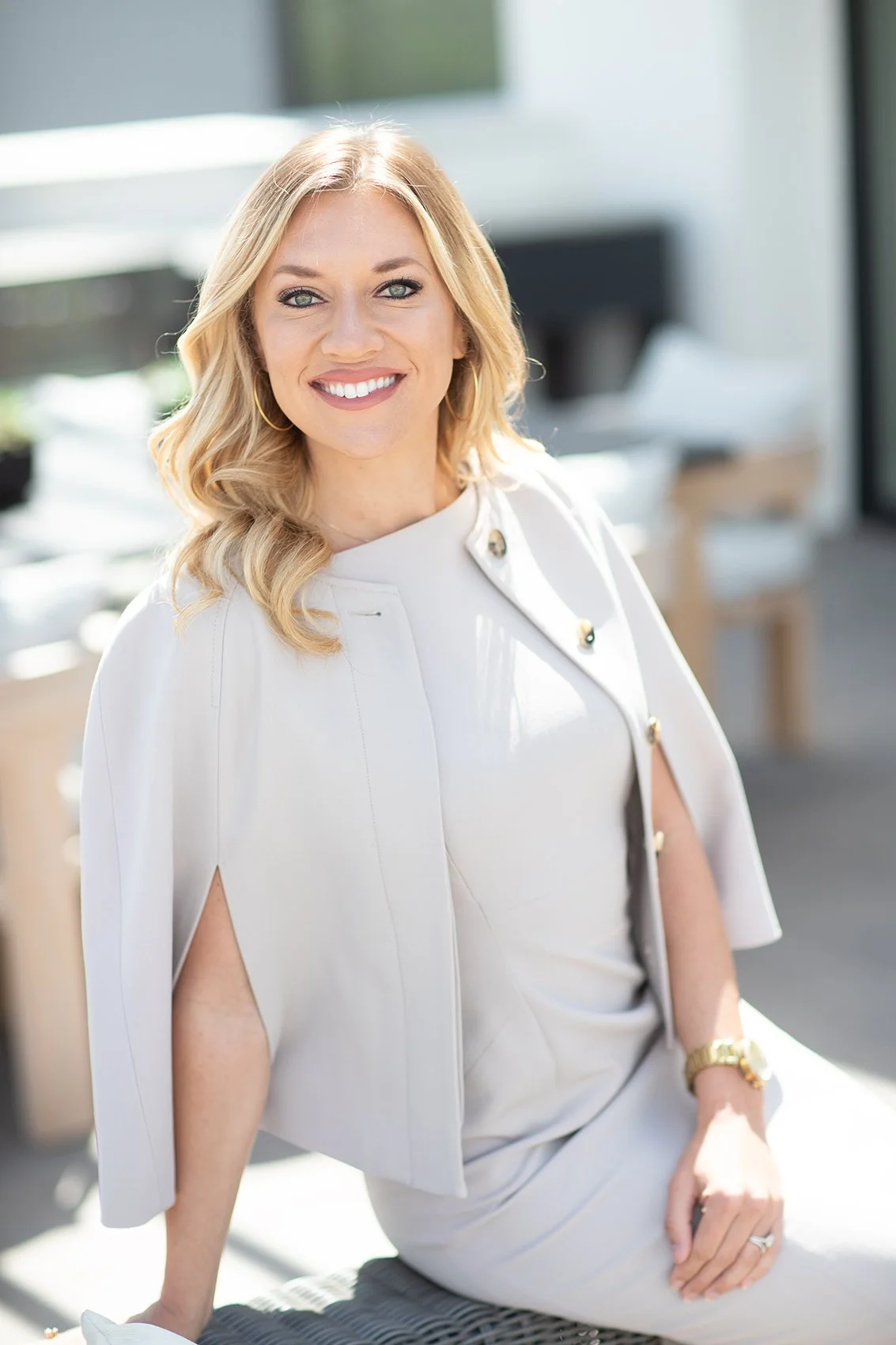 A woman with blonde hair and blue eyes smiling at the camera, wearing a white dress and a matching light jacket, sitting outdoors with a blurred background of a modern building.