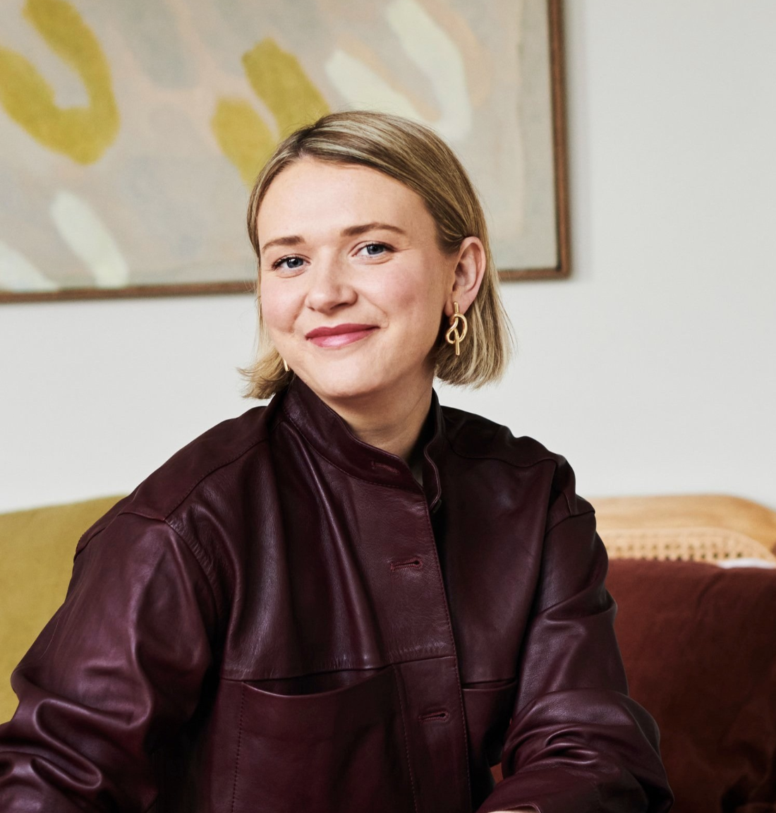 A young woman with blonde hair, blue eyes, and light skin, smiling, wearing a burgundy leather jacket and gold earrings, seated indoors with abstract art on the wall behind her.