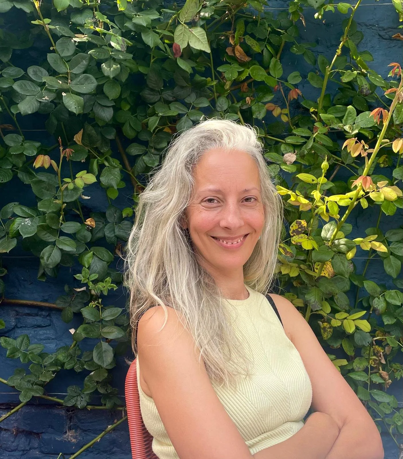 A smiling woman with long, curly gray hair posing outdoors in front of lush green foliage and a blue wall.
