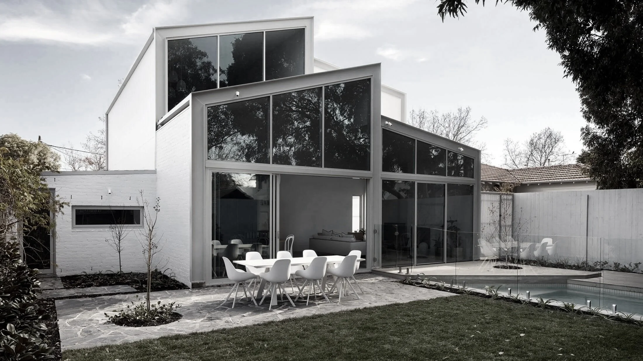 Modern white house with large glass windows and patio furniture, small backyard with trees and a pool, under cloudy sky.