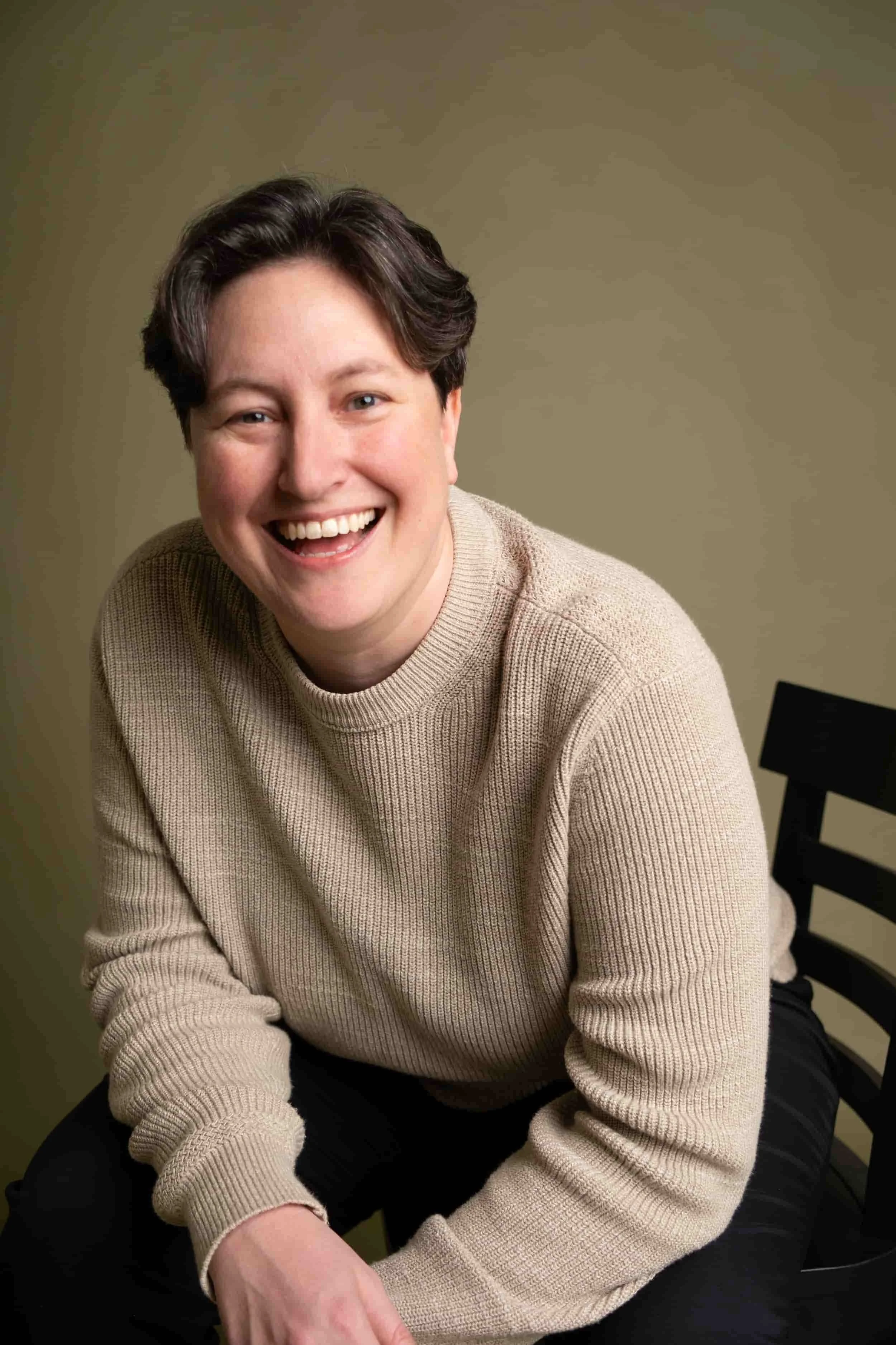 A woman with short brown hair, wearing a beige sweater, sits on a chair and smiles warmly at the camera against a plain background.