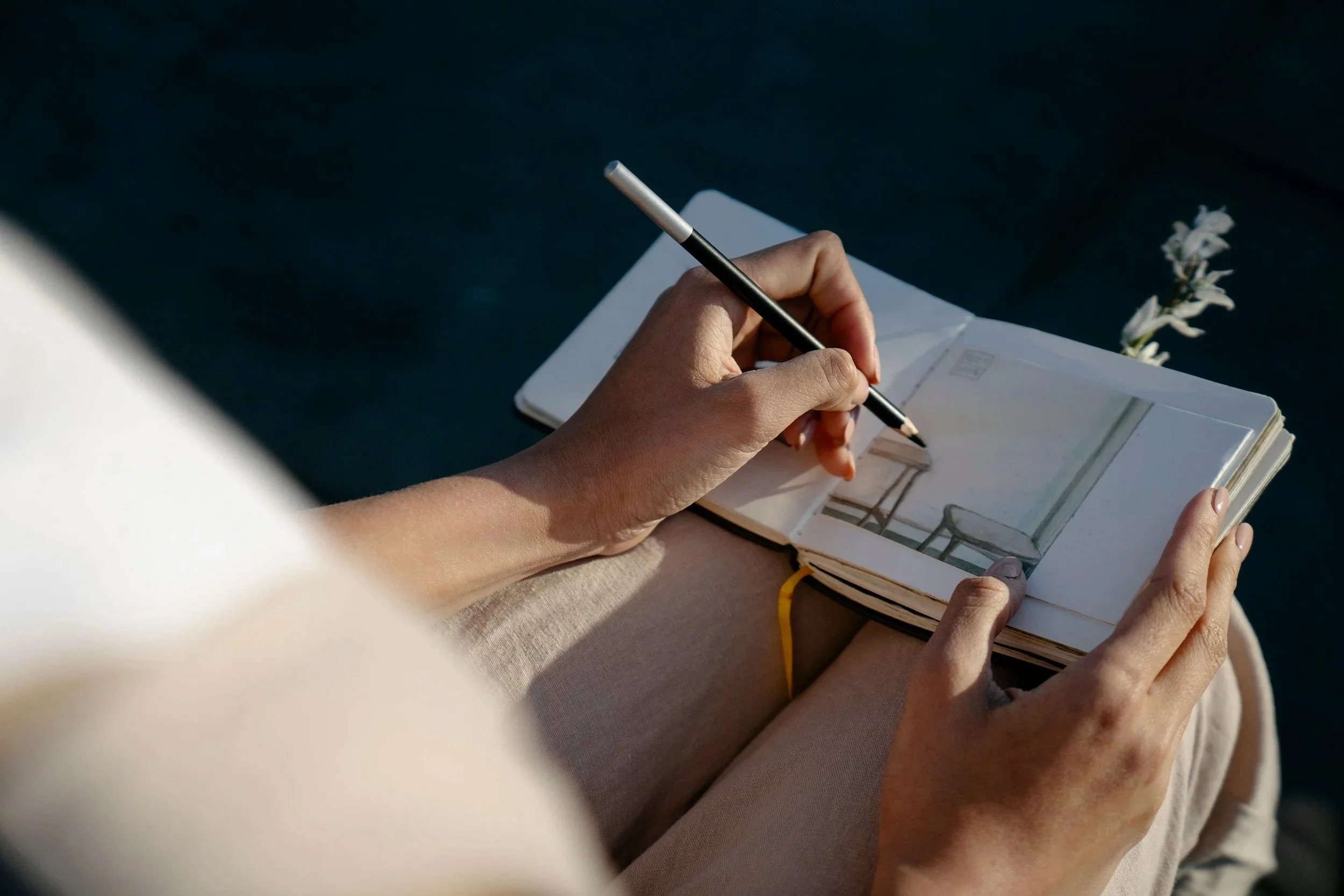 Person sitting outdoors, sketching and writing in a notebook with a pen, with a small flower tucked into the notebook.