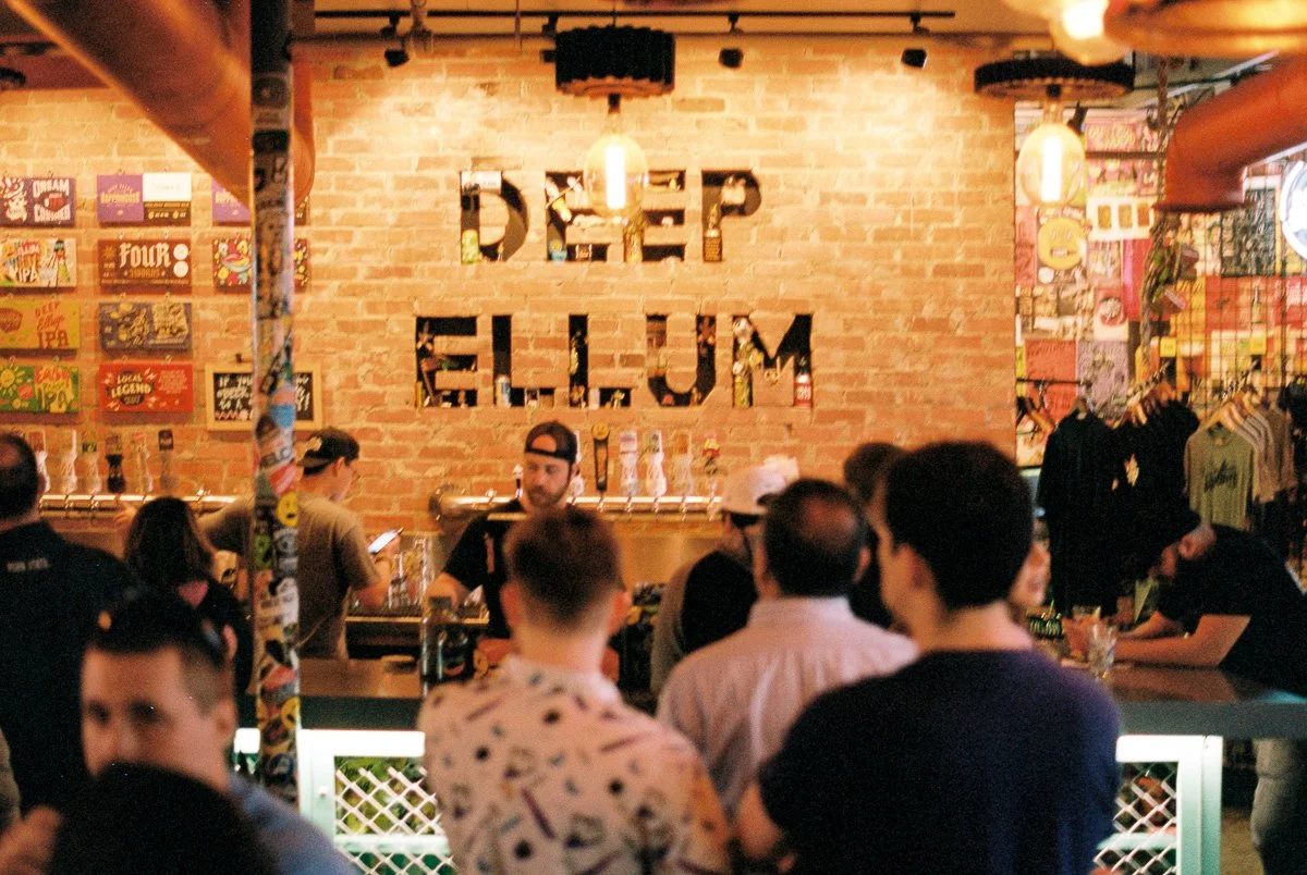People gathered inside a bar with a brick wall that displays the text "deep ellum" behind the bar area. Bar staff are preparing drinks, and colorful posters and clothing are visible in the decor.