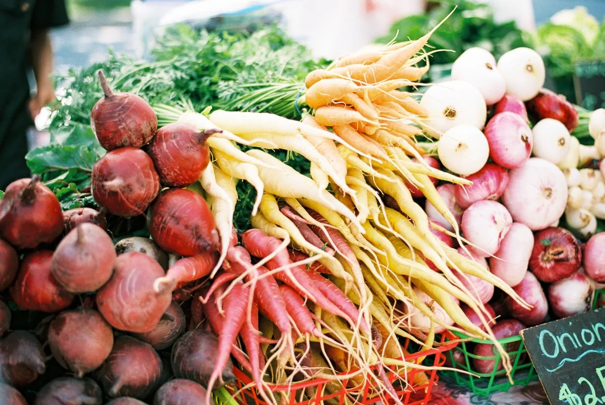 A display of colorful fresh vegetables at the farmer's market, including beets, yellow and pink onions, and carrots.