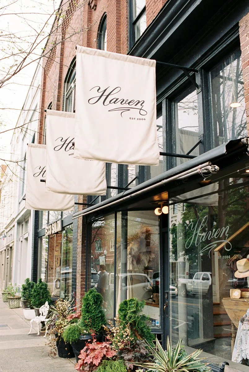 Street view of a boutique called Haven, with large windows, potted plants outside, and three hanging signs.