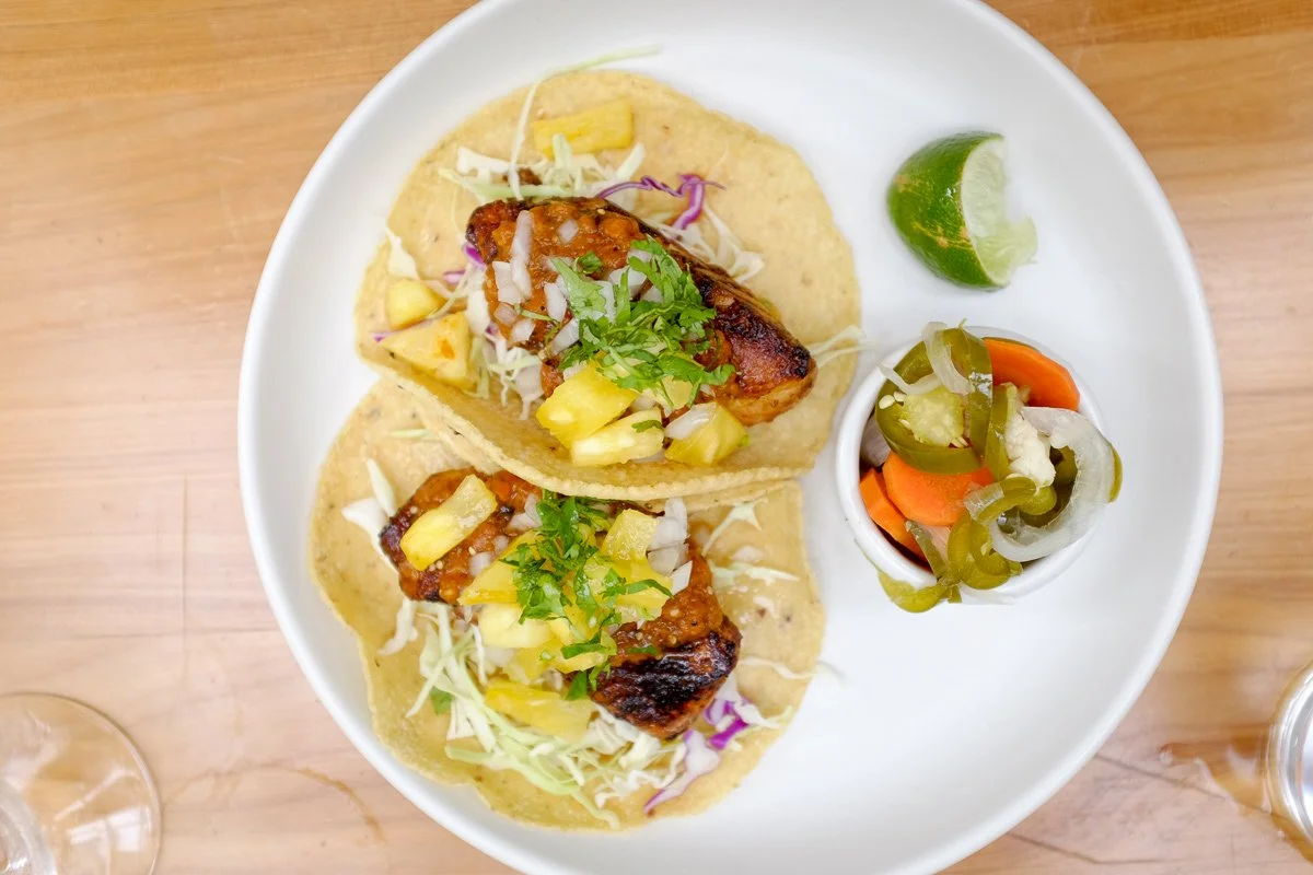 Two fish tacos with coleslaw, pineapple, and green herbs on a white plate, served with lime wedges and pickled jalapeños and carrots.