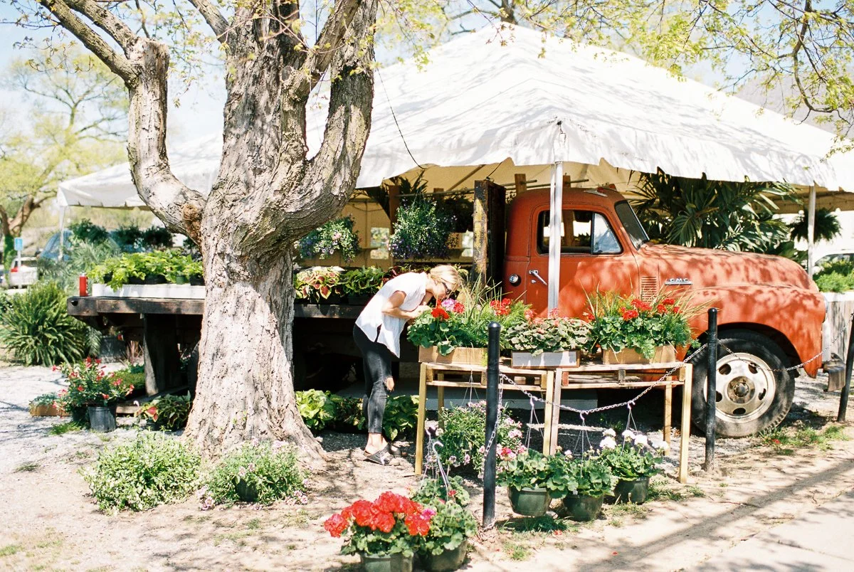 Ella browsing flowers at an outdoor flower stand next to a vintage rust-colored truck and a large tree under a white canopy.