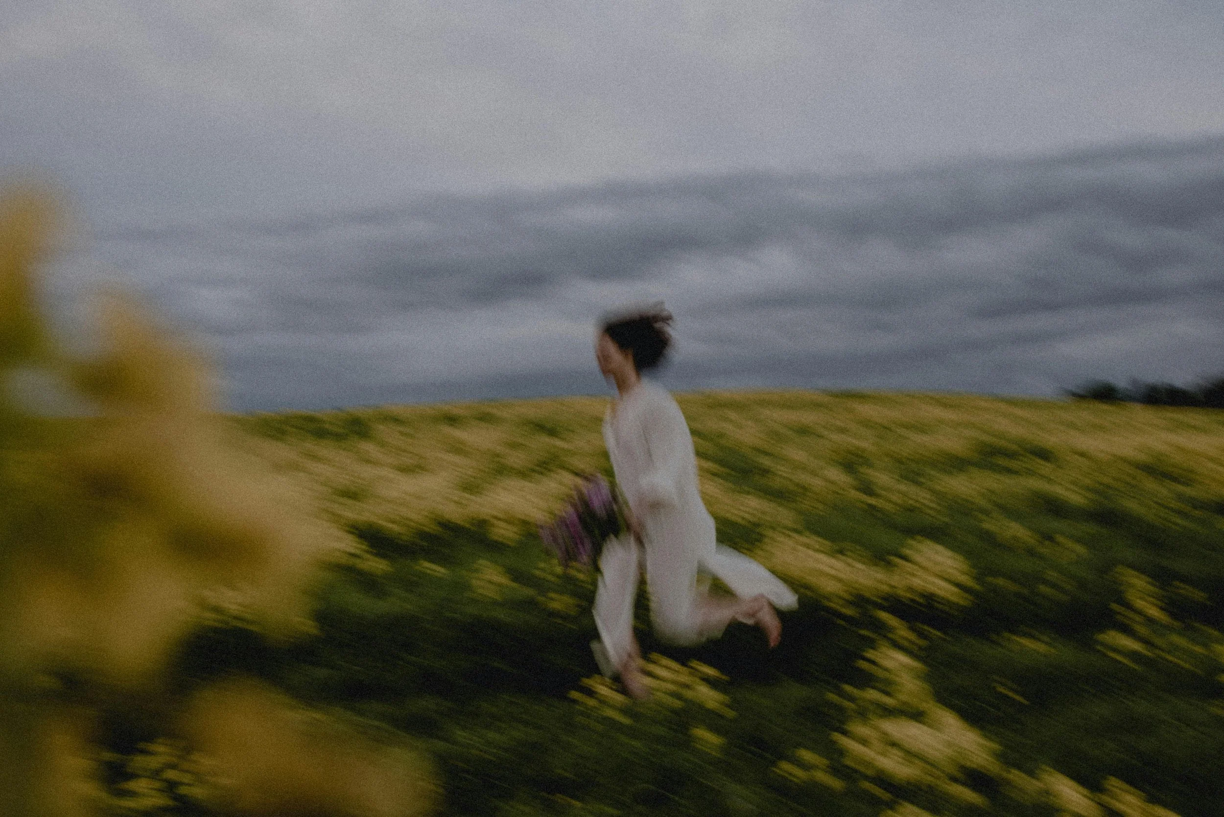 A person in white clothing running through a field of yellow flowers under a cloudy sky, appearing blurred due to motion.