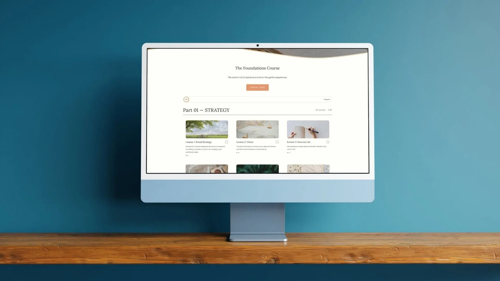 A computer monitor on a wooden desk displays an online course webpage titled 'The Foundations Course,' with a section called 'Part 01 – STRATEGY' featuring the first three lessons.