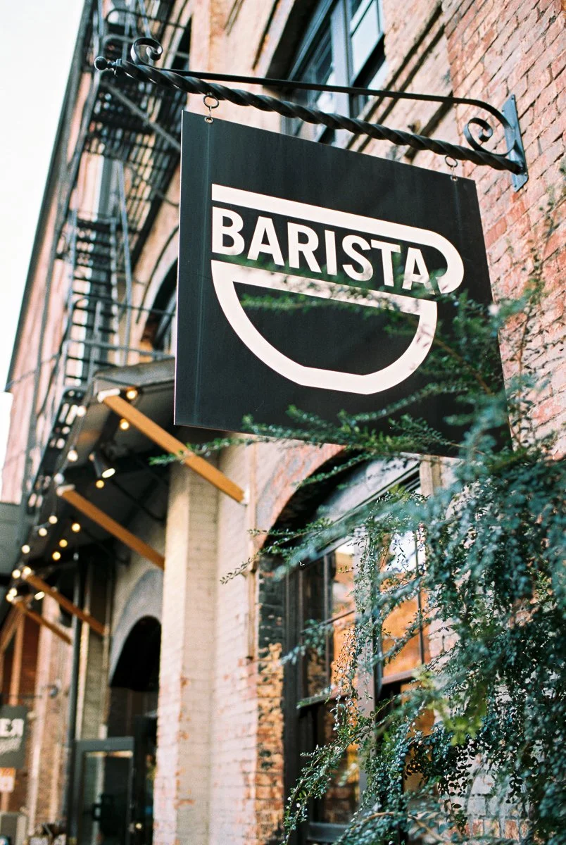 A black and white sign hanging outside a building reads 'Barista'. The building has a brick exterior with fire escape stairs and windows.