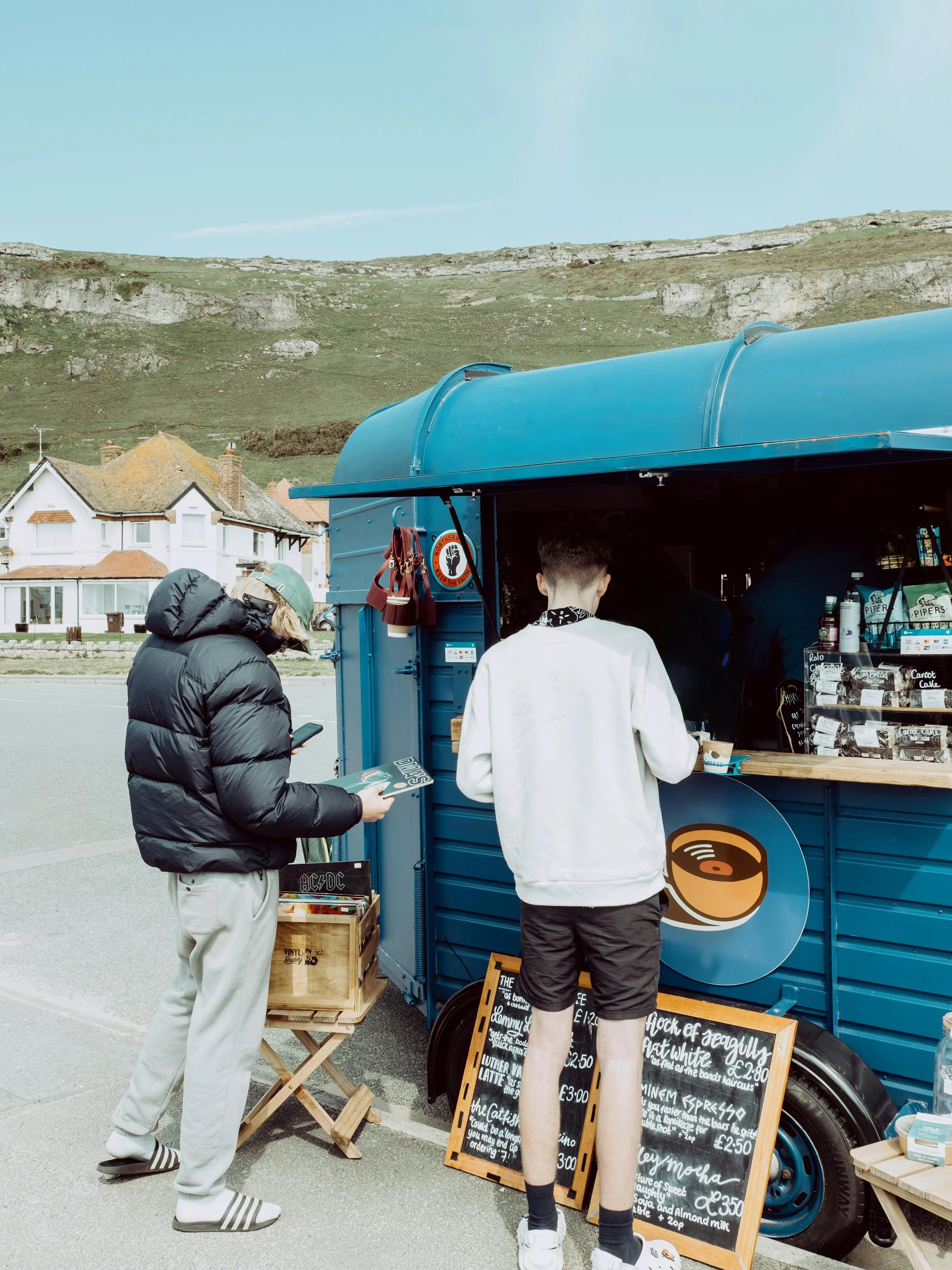 Two people buy coffee from a blue mobile coffee cart outdoors.