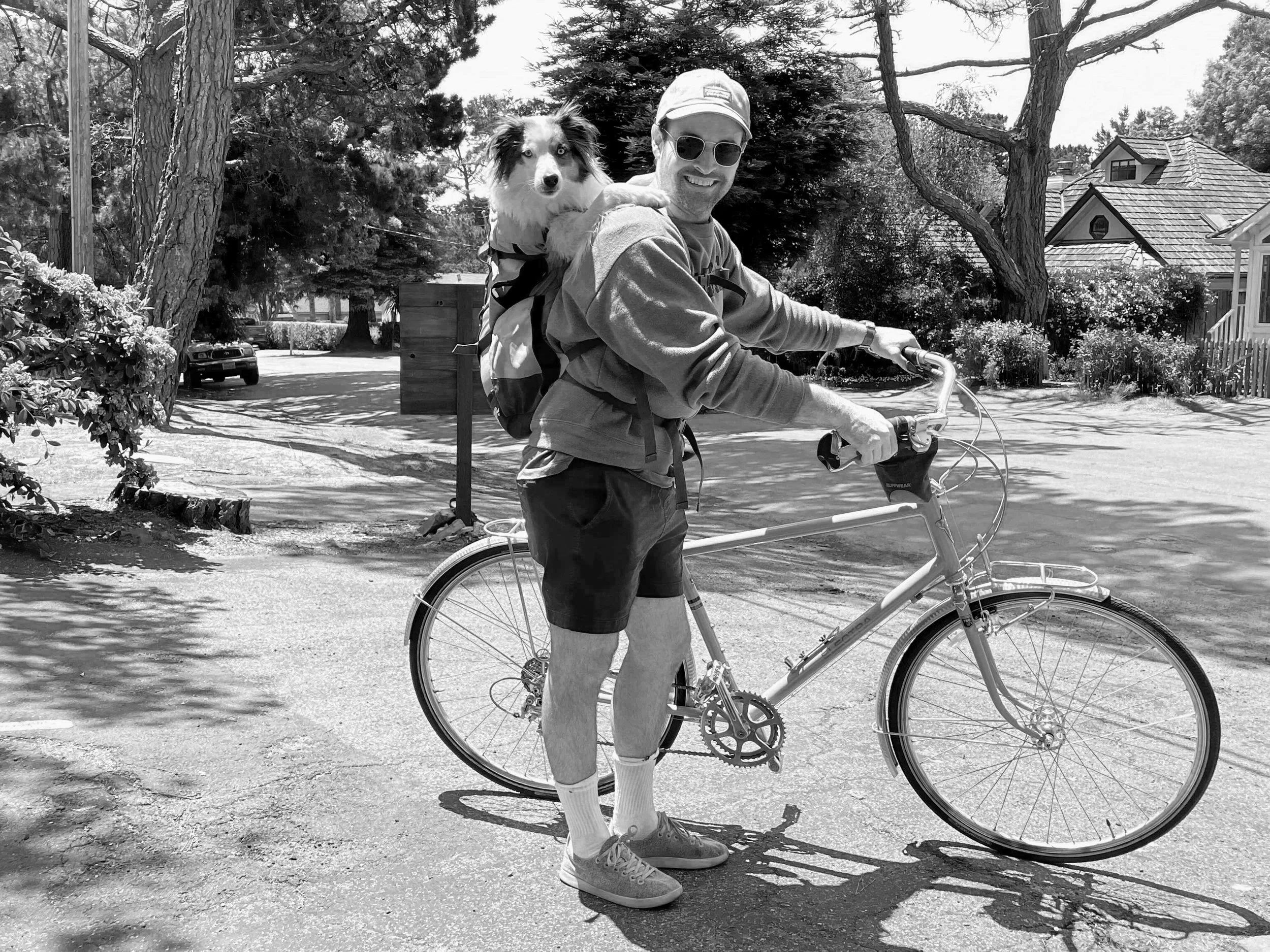 Ben riding a bicycle with a dog in a backpack on his back, smiling and wearing sunglasses, cap, shorts, and sneakers on a village street with trees and houses.