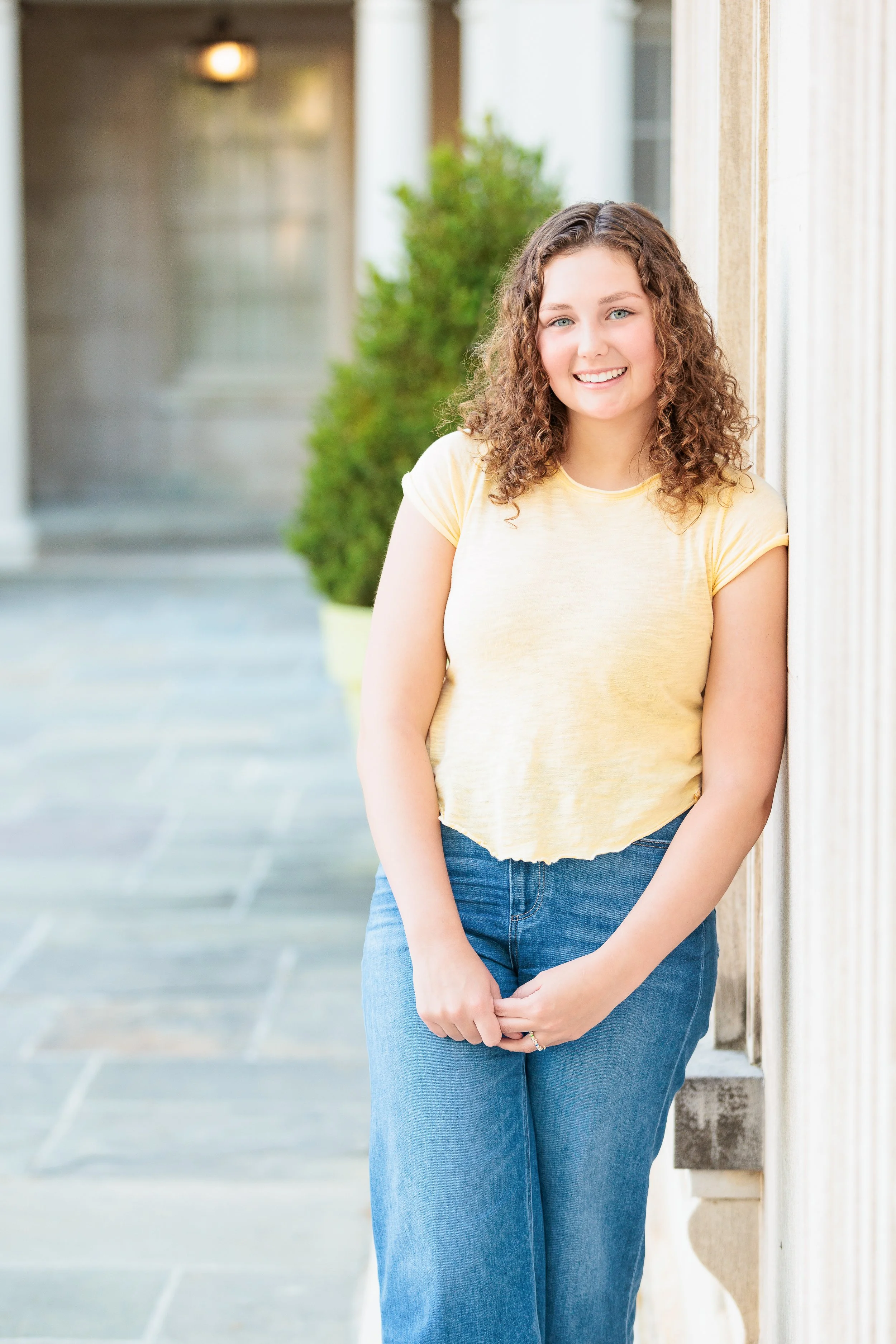 Young woman with curly hair smiling and leaning against a wall outdoors.