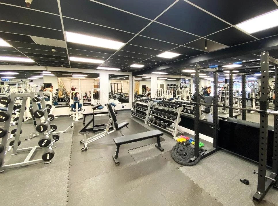 Interior view of a gym with various workout equipment, including dumbbells, weight racks, benches, and a lifting cage, with a mirror on the wall.