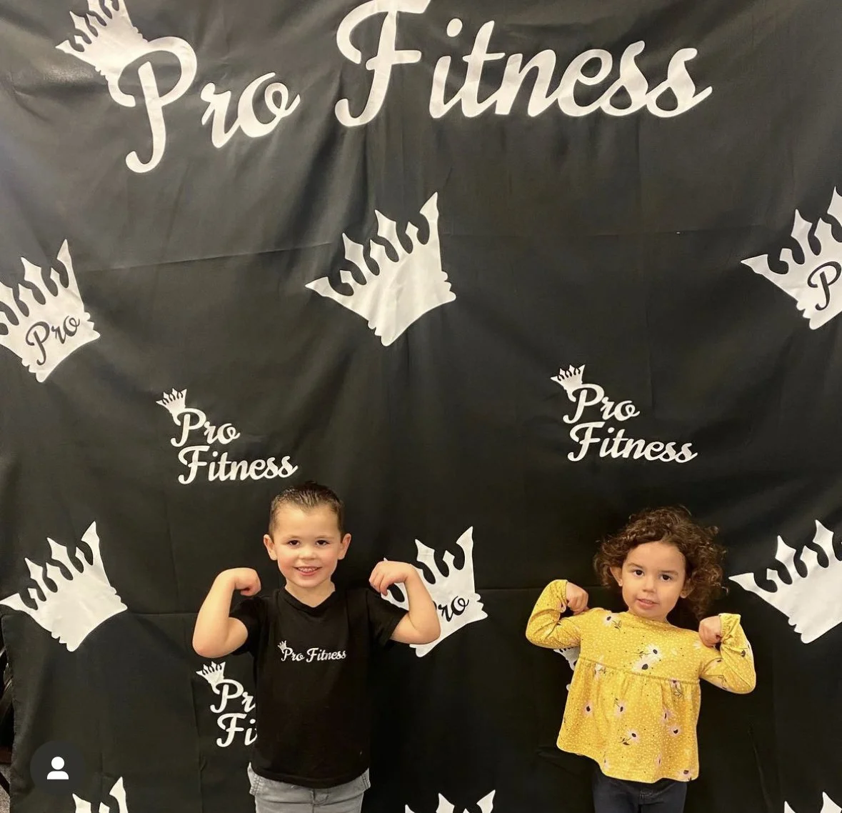 Two children posing in front of a black backdrop with white 'Pro Fitness' logos and crowns. The boy on the left is smiling, wearing a black t-shirt with the 'Pro Fitness' logo, and flexing his arms. The girl on the right, with curly hair and a seriou