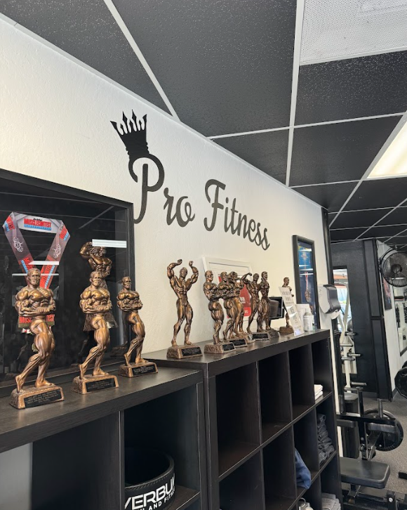 Interior of a gym with a wall that says "Pro Fitness" and several bodybuilding trophies displayed on a black shelf below the sign.