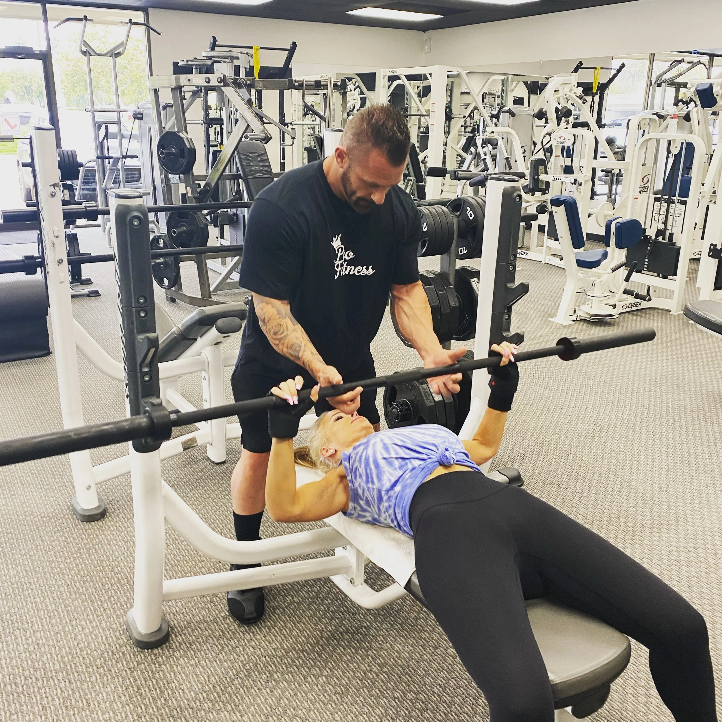 A woman lying on a bench lifting a barbell with a trainer standing over her in a gym.