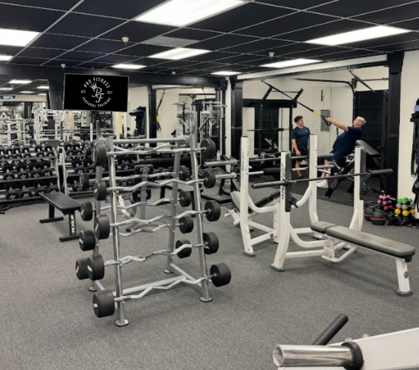 Interior of a gym with various workout equipment, including weight racks, benches, and machines. Two people are working out near a cable machine, with one person assisting or coaching the other.