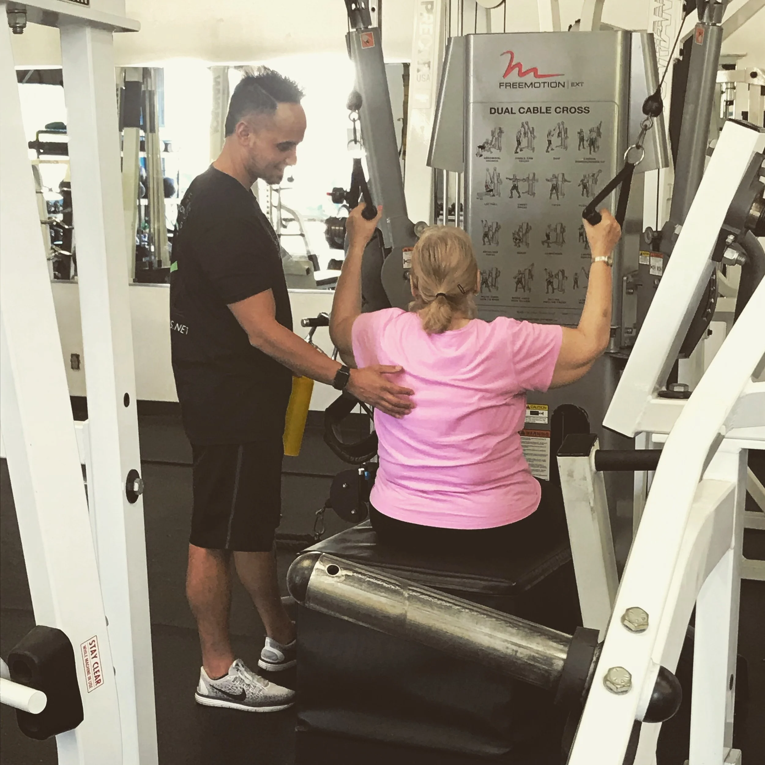 A woman in a pink shirt is seated on a black exercise bench, lifting weights with her arms, while a trainer in a black shirt assists her at a gym. The trainer is helping guide her exercise at a machine labeled 'Dual Cable Cross'.