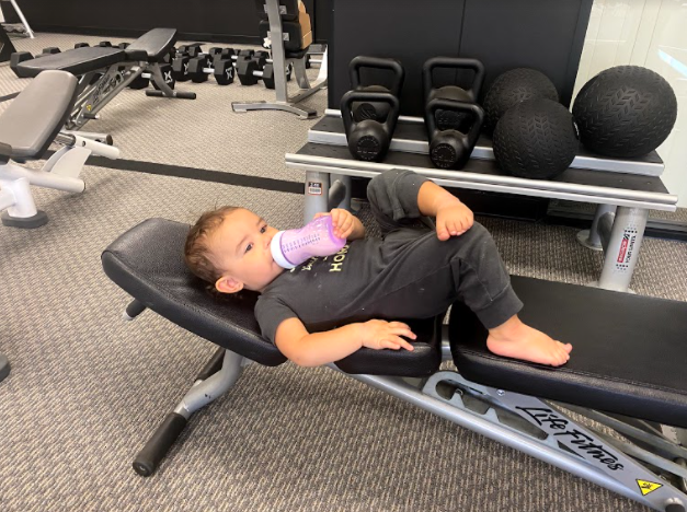 A young boy lying on a workout bench in a gym, drinking from a pink water bottle.