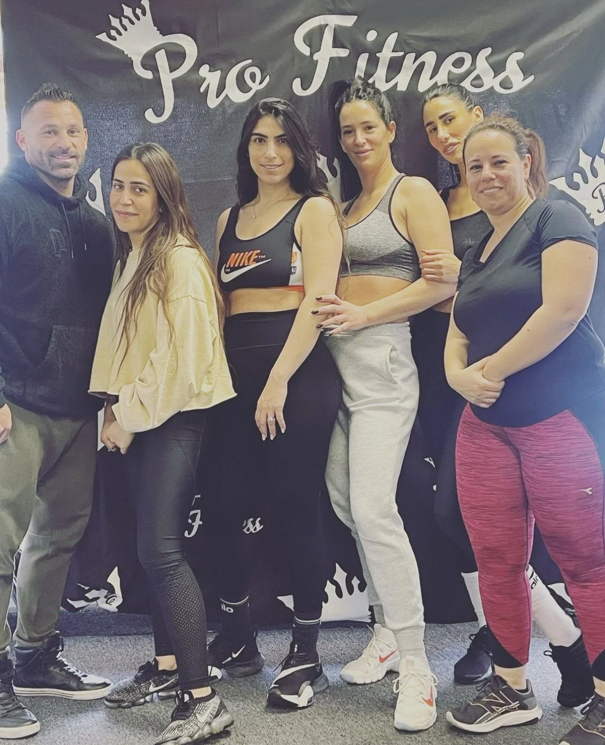 Group of six people, five women and one man, standing together at a gym with a "Pro Fitness" banner in the background.