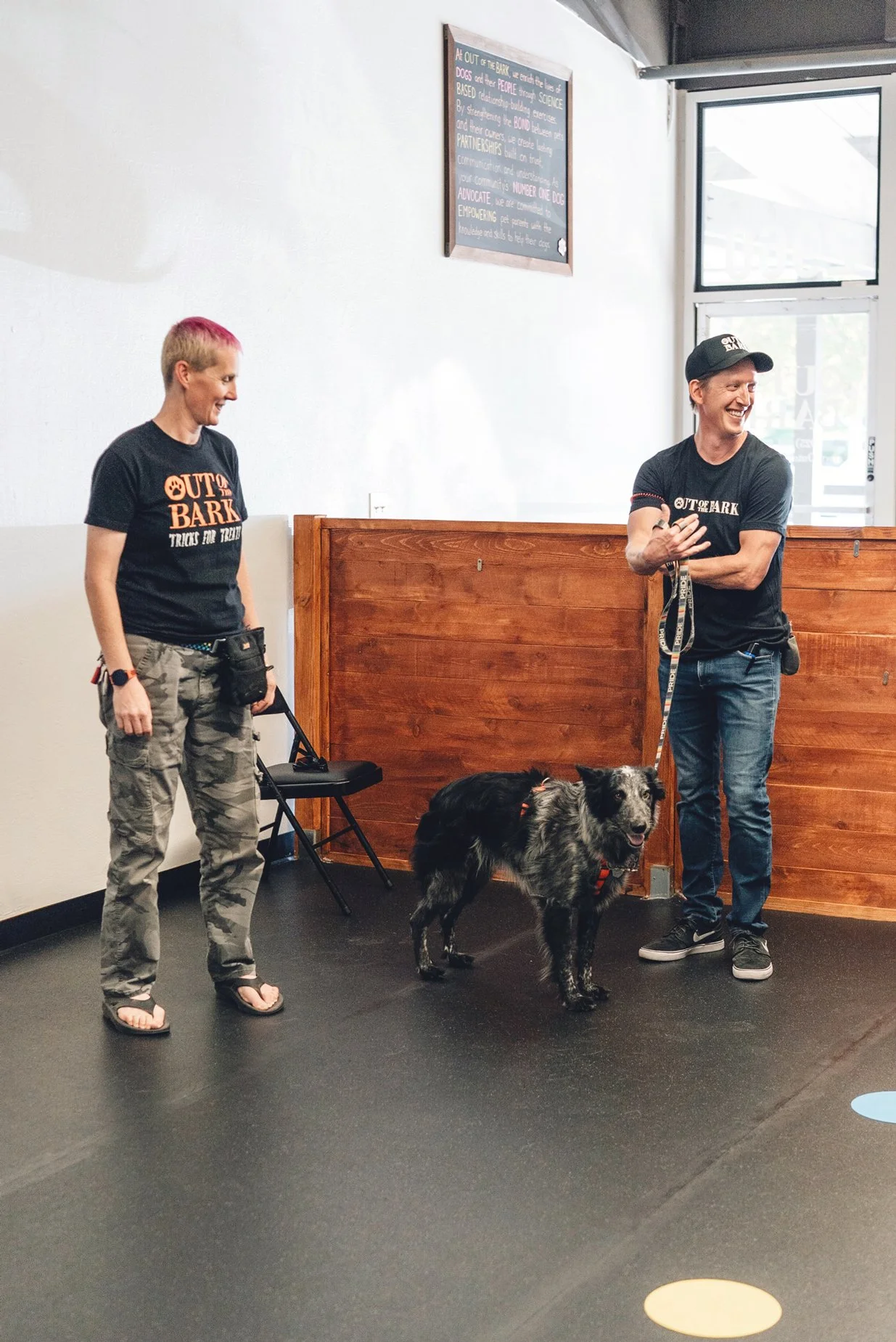 Two people smiling at each other during a dog training session with a black and gray dog on a leash inside a room with black flooring and wooden wall paneling.