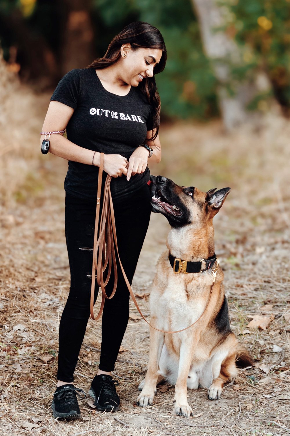 A woman holding a long leash while a German shepherd sits at her side outdoors, looking up at her face.