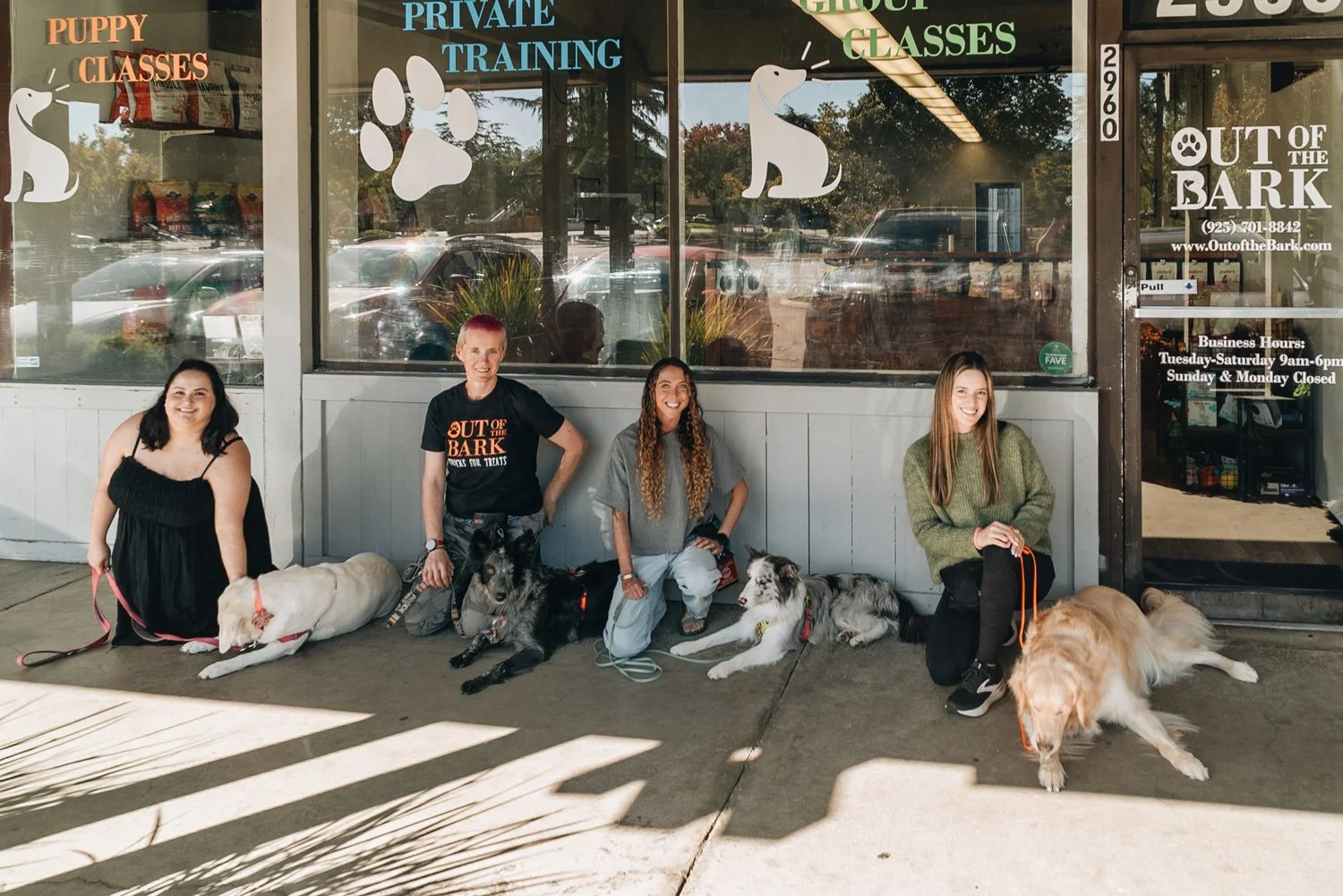 Four women sitting in front of 'Out of the Bark' training facility with their dogs, smiling and posing for the photo. The store storefront features paw print and dog images, and has signs for puppy classes and private training.