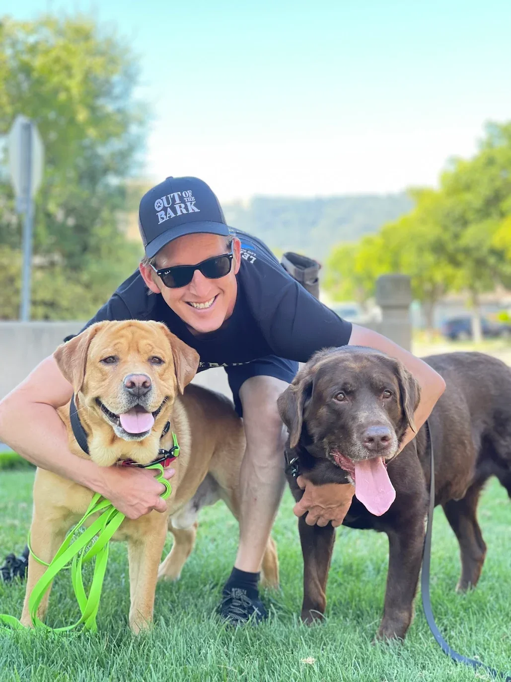 A man wearing sunglasses, a black cap reading 'OUT OF THE BARK,' and black T-shirt, crouching on grass, holding two dogs. One dog is tan with a green leash, and the other is dark brown with a pink tongue hanging out. The background features trees and a partly cloudy sky.