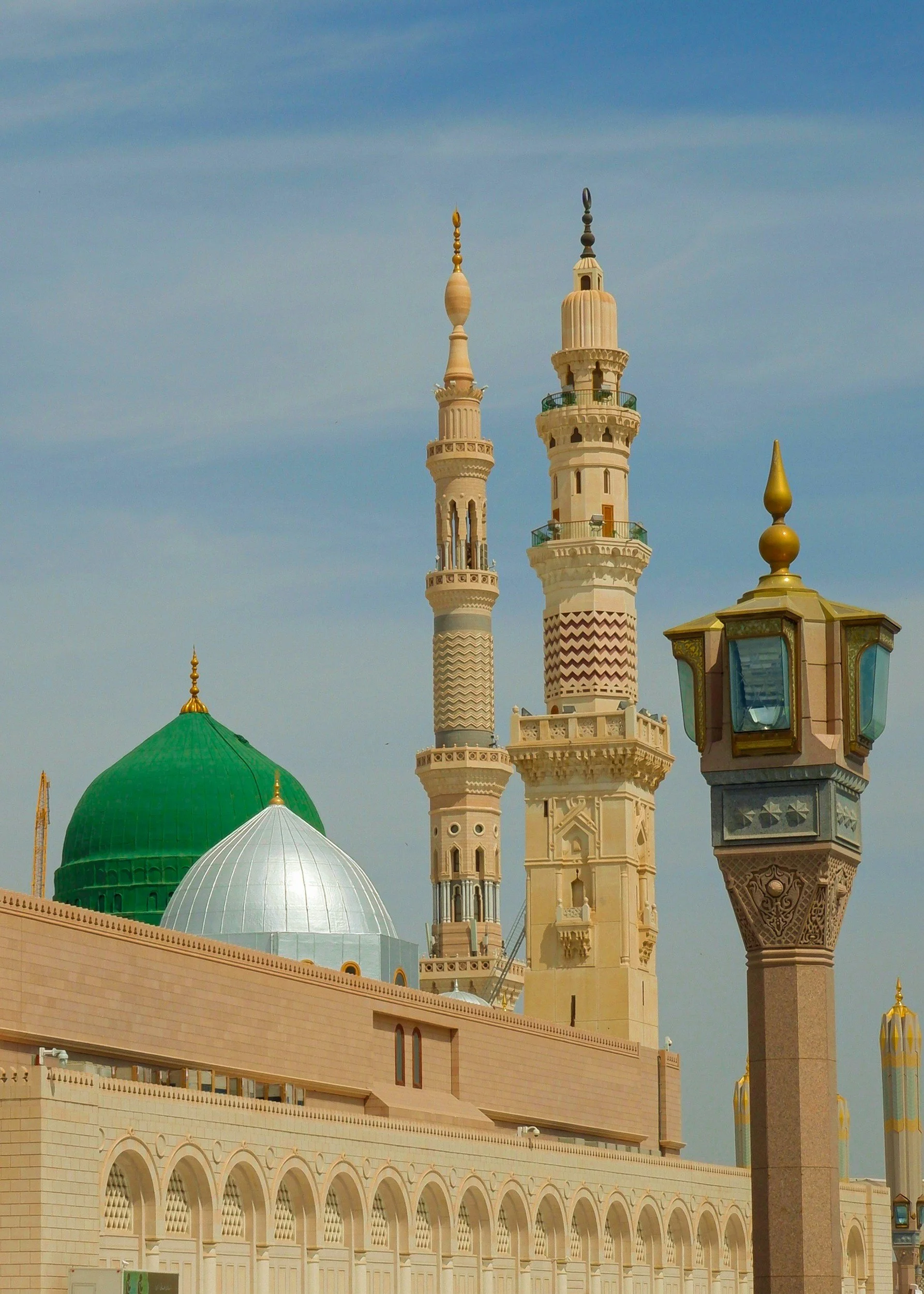 View of a mosque with green, white, and beige domes and two tall minarets, with a decorative street lamp in the foreground, under a blue sky.