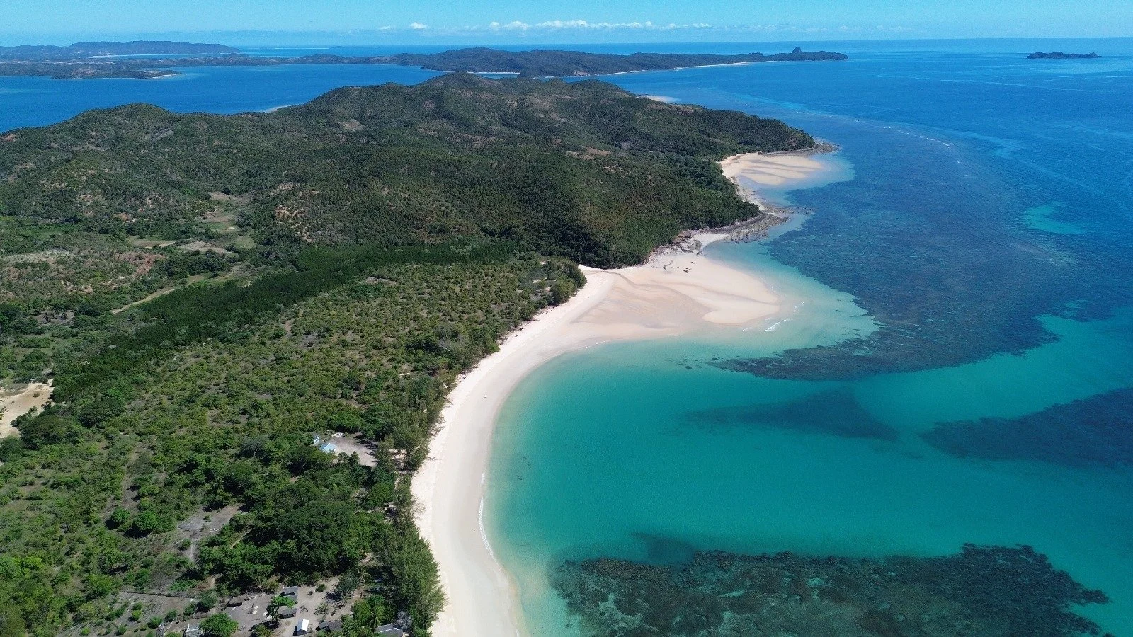Luftaufnahme eines weißen Sandstrandes an einer grünen Küstenregion mit türkisfarbenem Wasser, umgeben von Wald und hügeligem Land, im Hintergrund das offene Meer