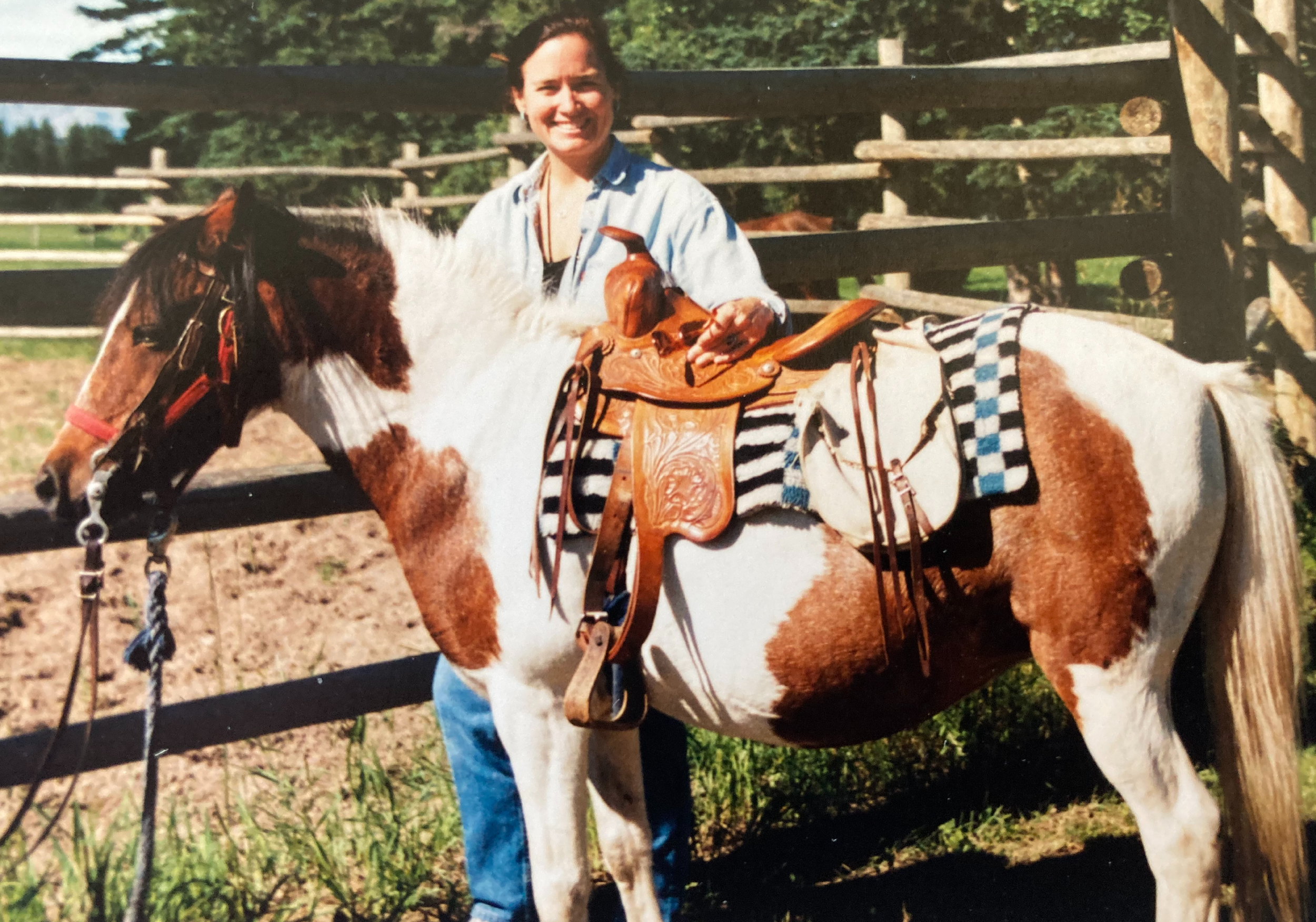 A woman with dark hair smiling while sitting on a white and brown horse with a saddle, in an outdoor paddock with a wooden fence and trees in the background.